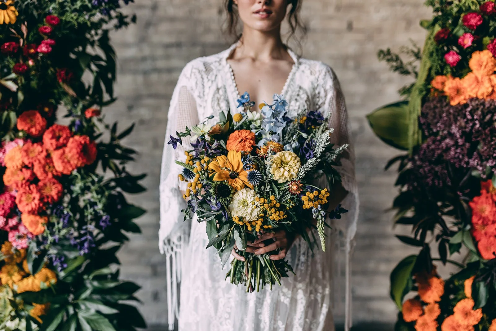 A woman in a white lace dress holds a large colorful bouquet of flowers. She is standing between two large flower arrangements against a brick wall.
