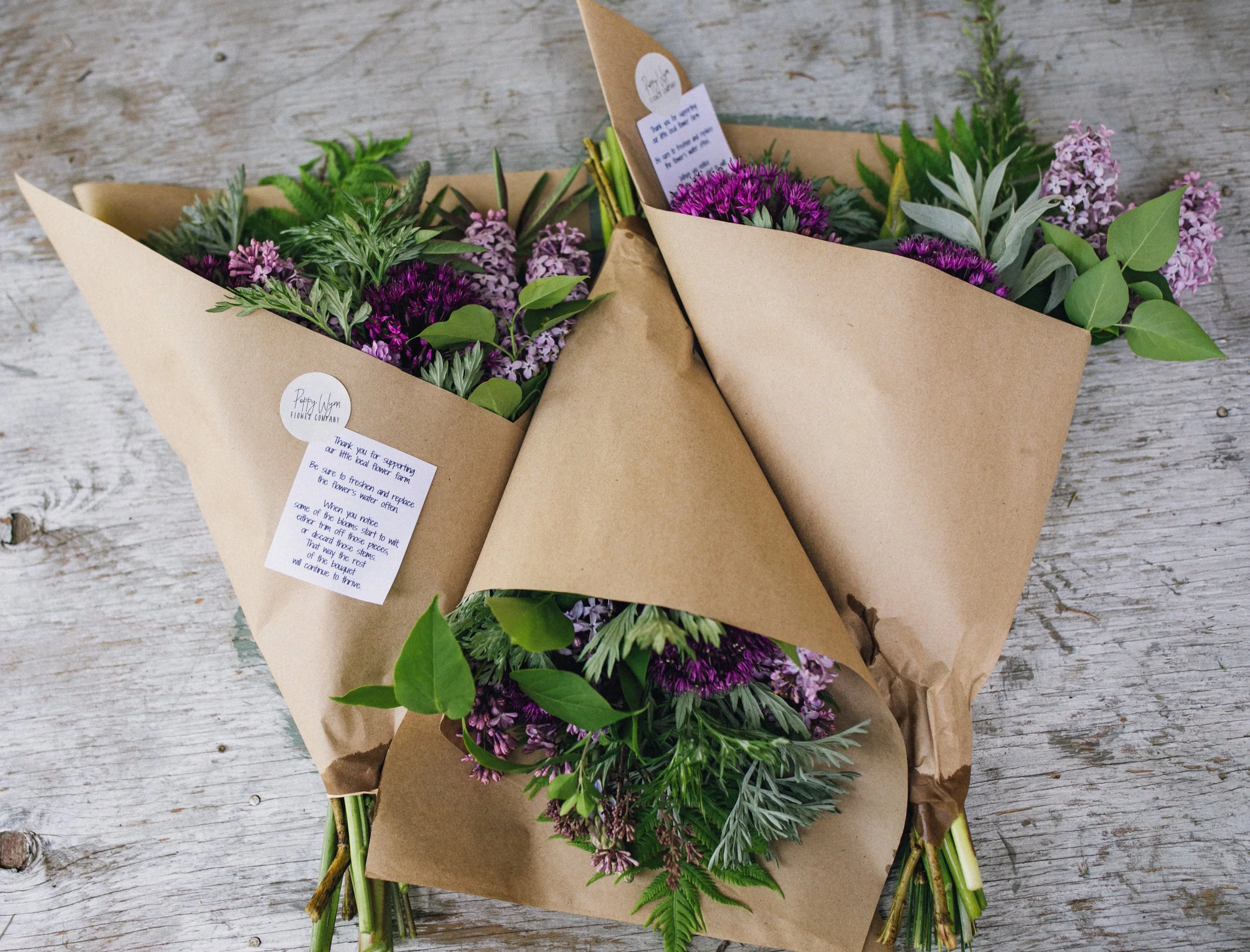 Two brown paper bouquets wrapped around purple and pink flowers and green leaves on a wooden surface.