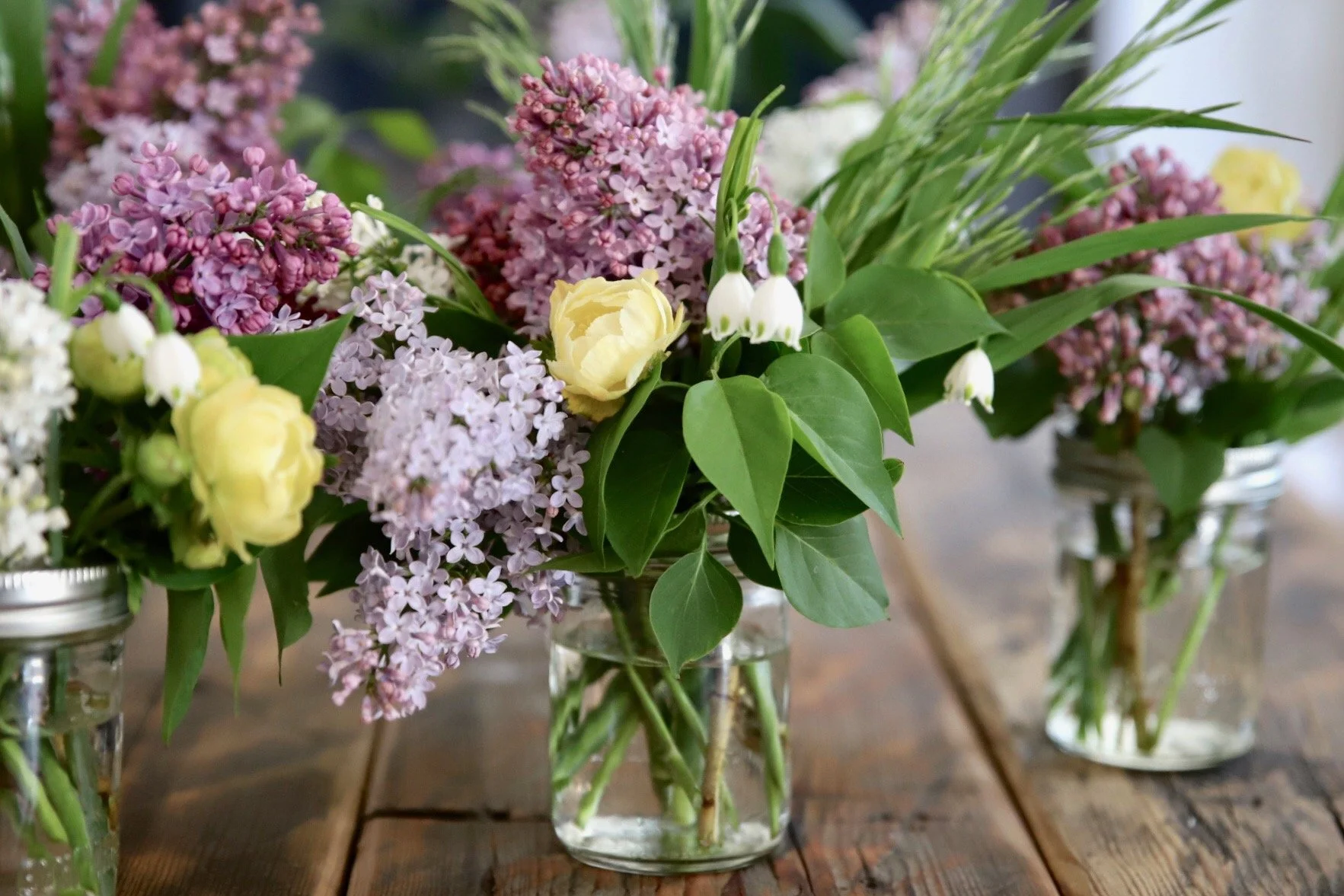 Several glass jars with flowers including lilacs, roses, and other greenery on a wooden table.