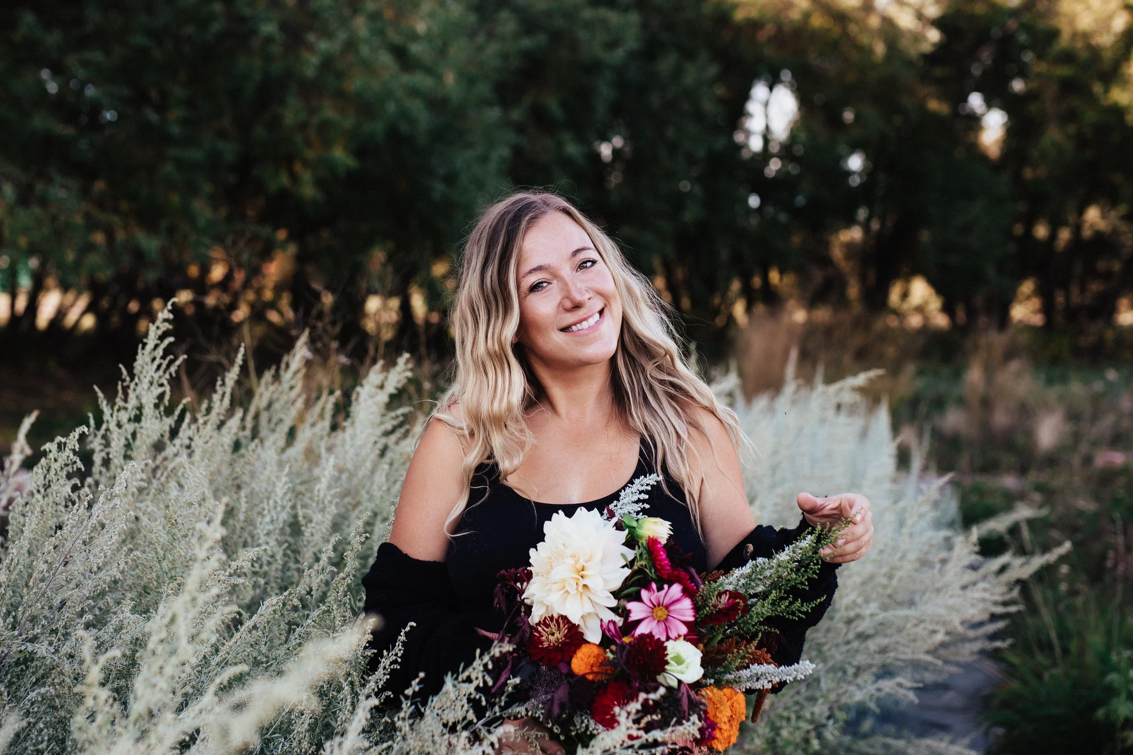 A woman with long wavy blonde hair smiling outdoors, holding a large colorful bouquet of flowers, with a background of trees and dry grass.