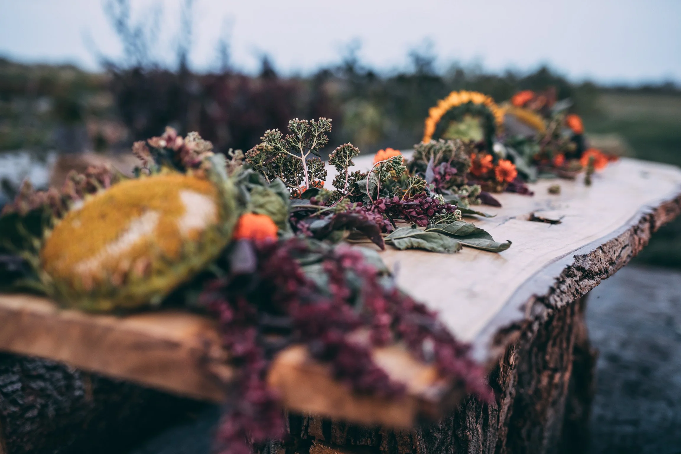 Close-up view of a decorative arrangement of leaves, flowers, and natural materials on a wooden surface outdoors, with blurred trees and sky in the background.