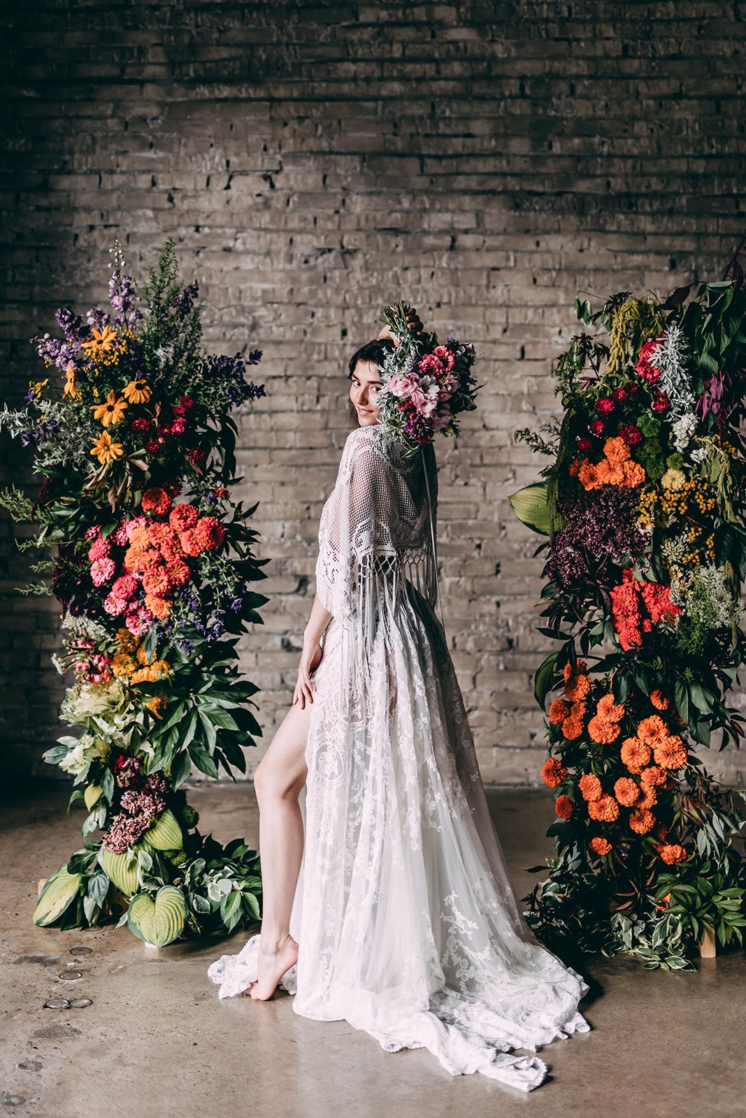 A woman in a sheer, lace, white dress and floral headpiece standing between two large floral arrangements against a brick wall.