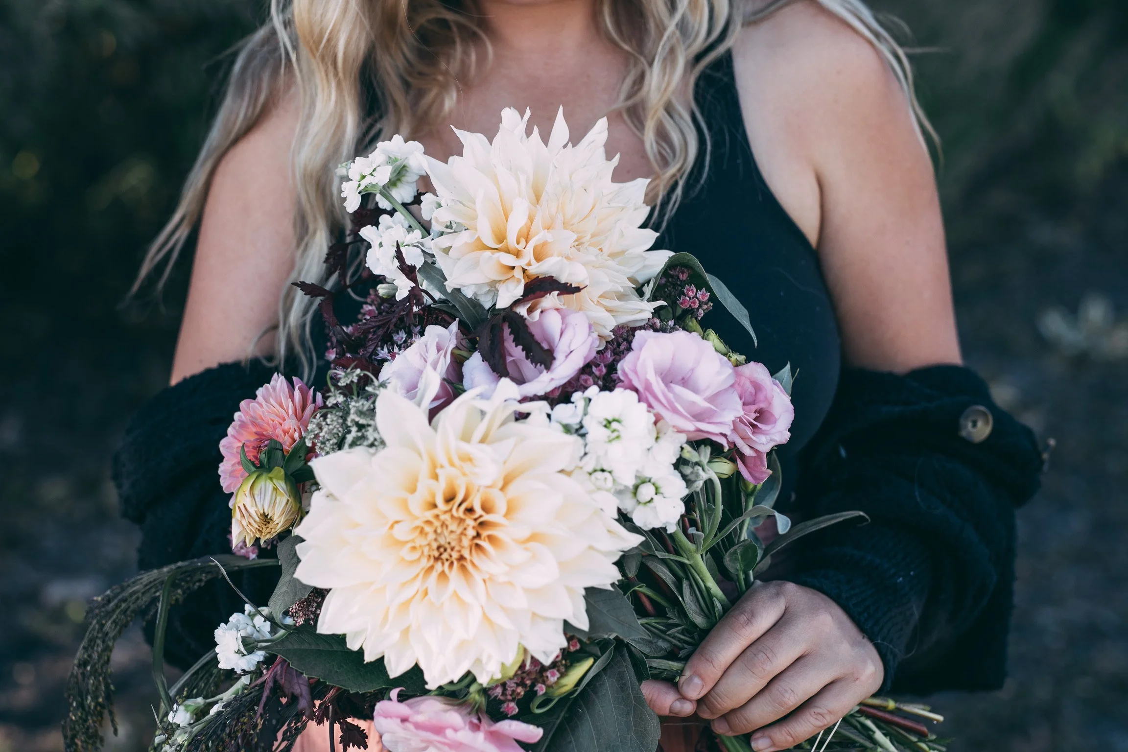 Person holding a large bouquet of mixed flowers including dahlias, roses, and other blossoms in soft pinks and creams.