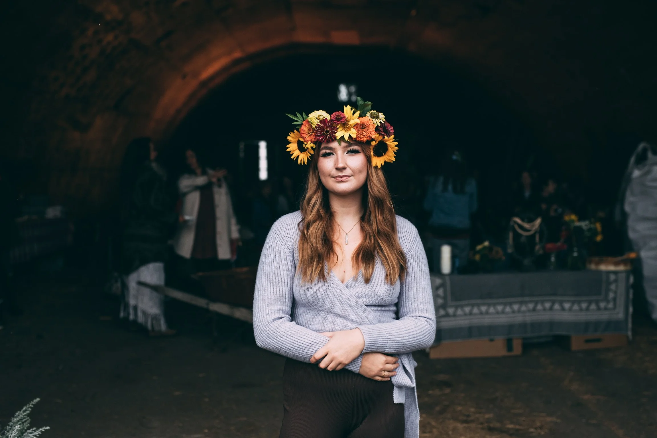 A woman with long red hair, wearing a sunflower crown, standing with arms crossed in front of an outdoor market stall.
