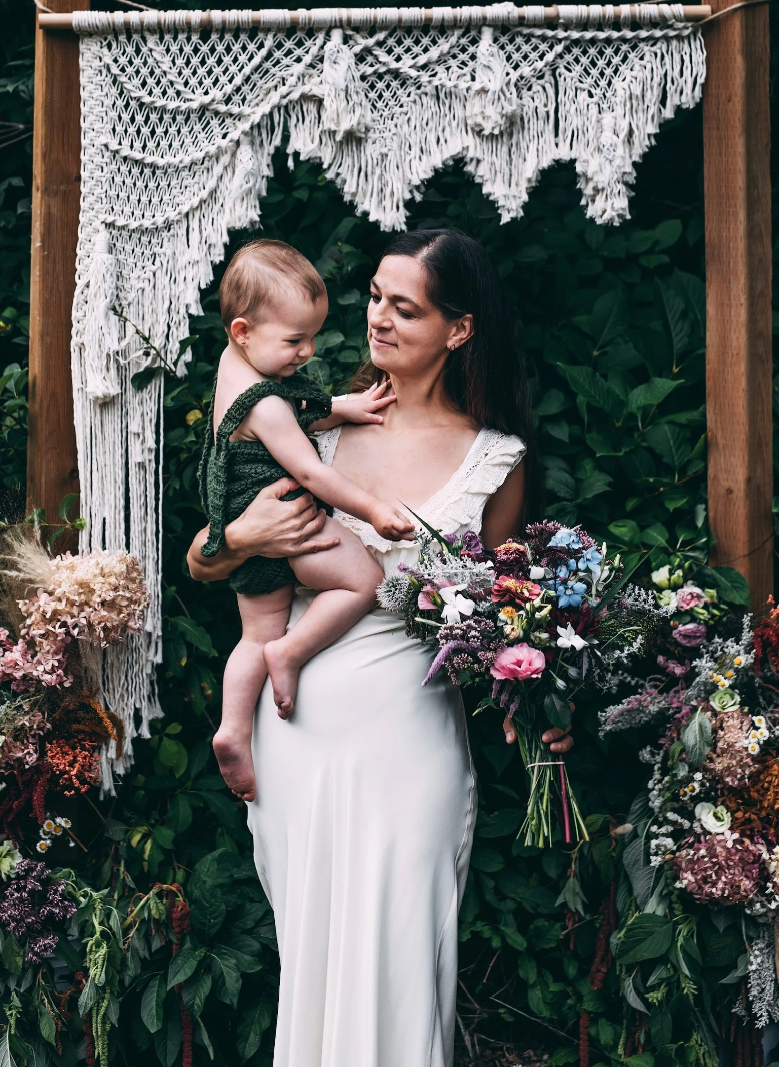 A woman in a cream-colored dress holds a young child in a dark green knit outfit amidst lush greenery and colorful flowers, with a white macrame decoration hanging above.