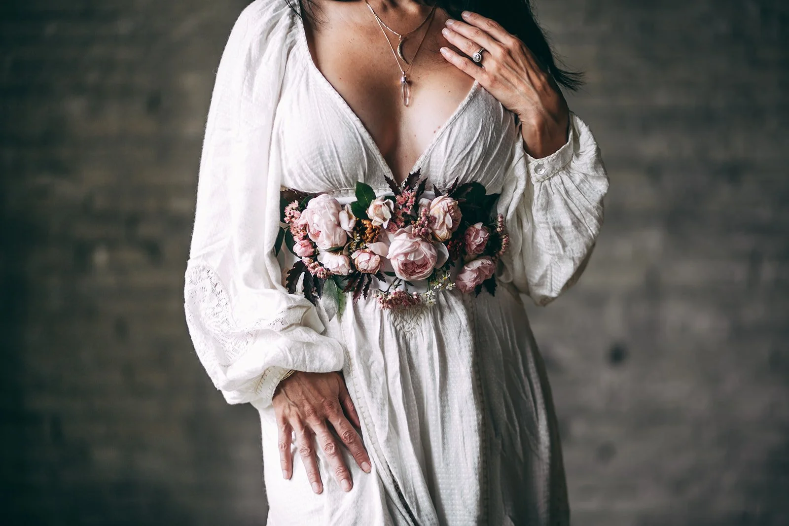 A woman in a vintage white dress with lace details is holding a bouquet of pink and white flowers, with her hand on her chest and waist, against a dark, blurred background.