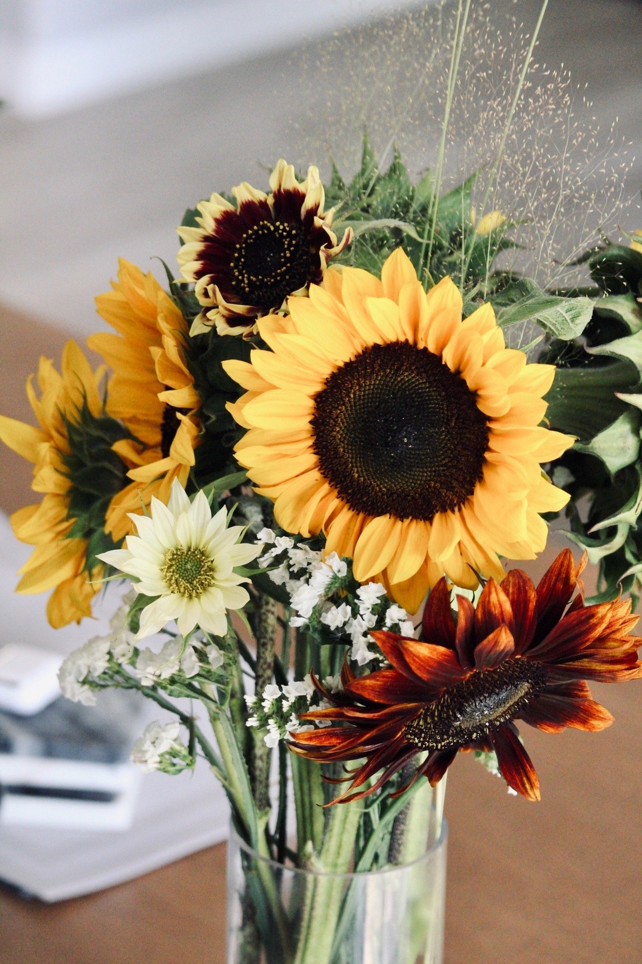 A bouquet of various colorful sunflowers and small white flowers in a clear glass vase on a wooden surface.