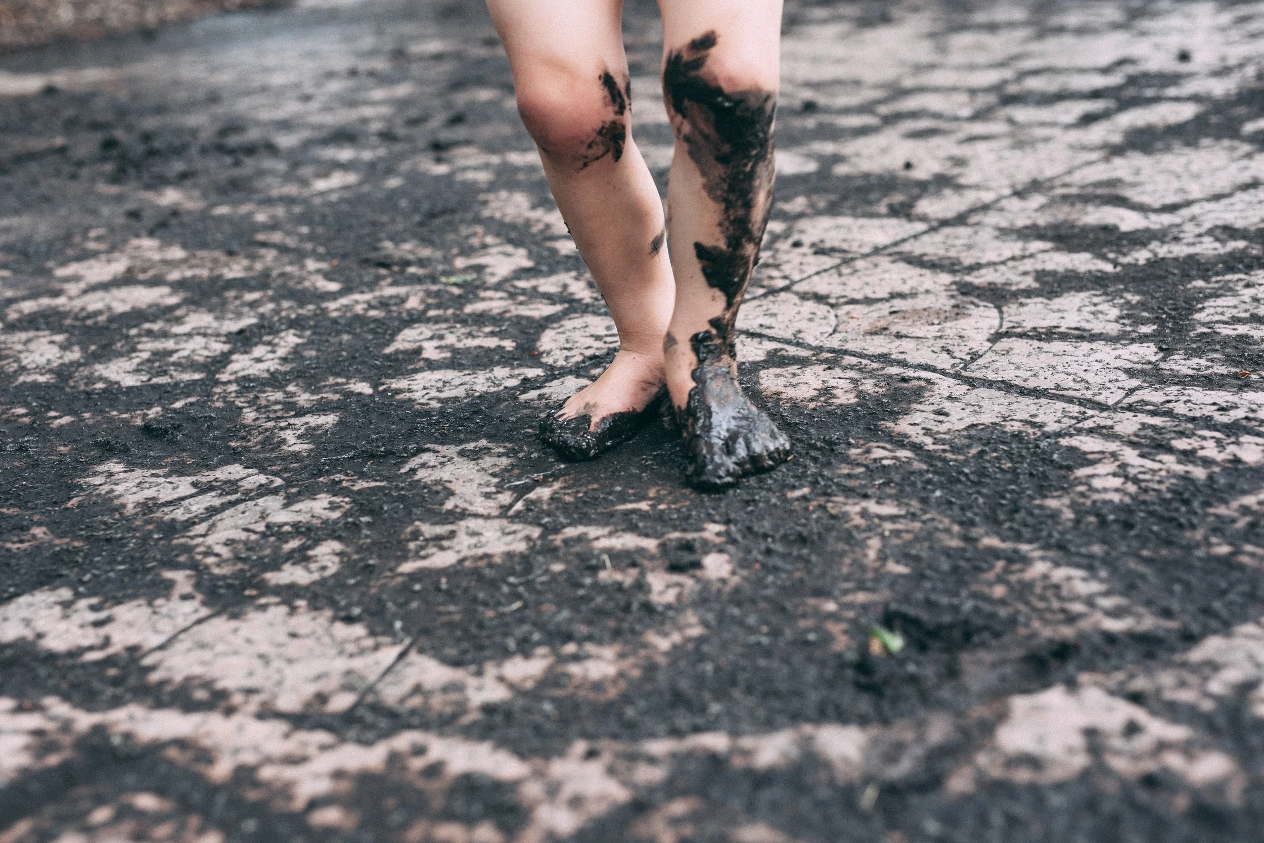 Bare legs and feet covered in mud walking on a cracked, muddy surface.