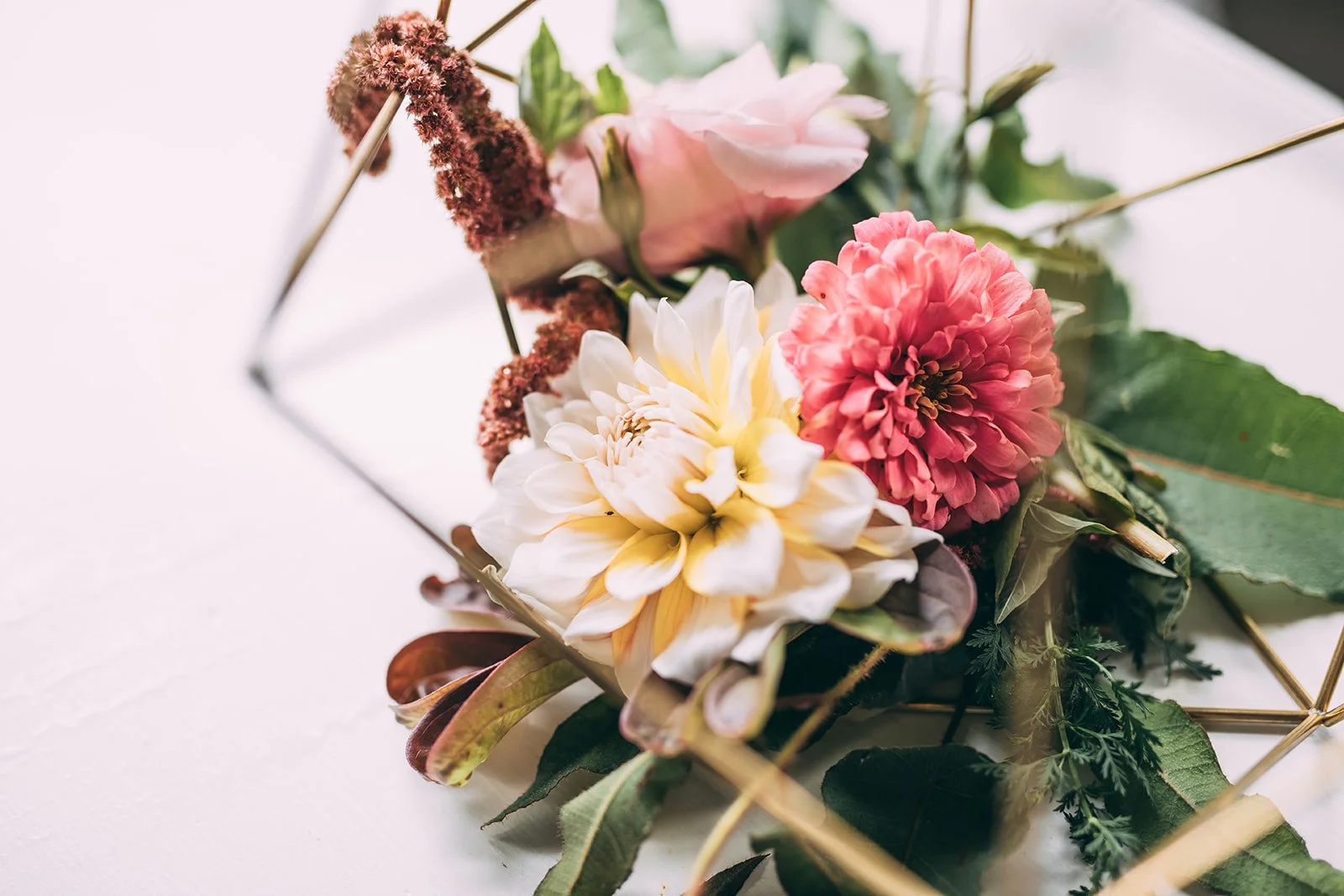 Close-up of mixed fresh flowers, including a pink rose, a white dahlia, and a pink carnation, surrounded by green leaves, on a white surface.