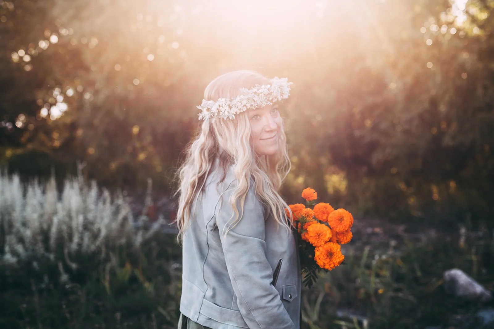 A woman with long, wavy blonde hair wearing a floral crown and a gray jacket, holding a bouquet of orange marigolds, standing outdoors during sunset with trees in the background.