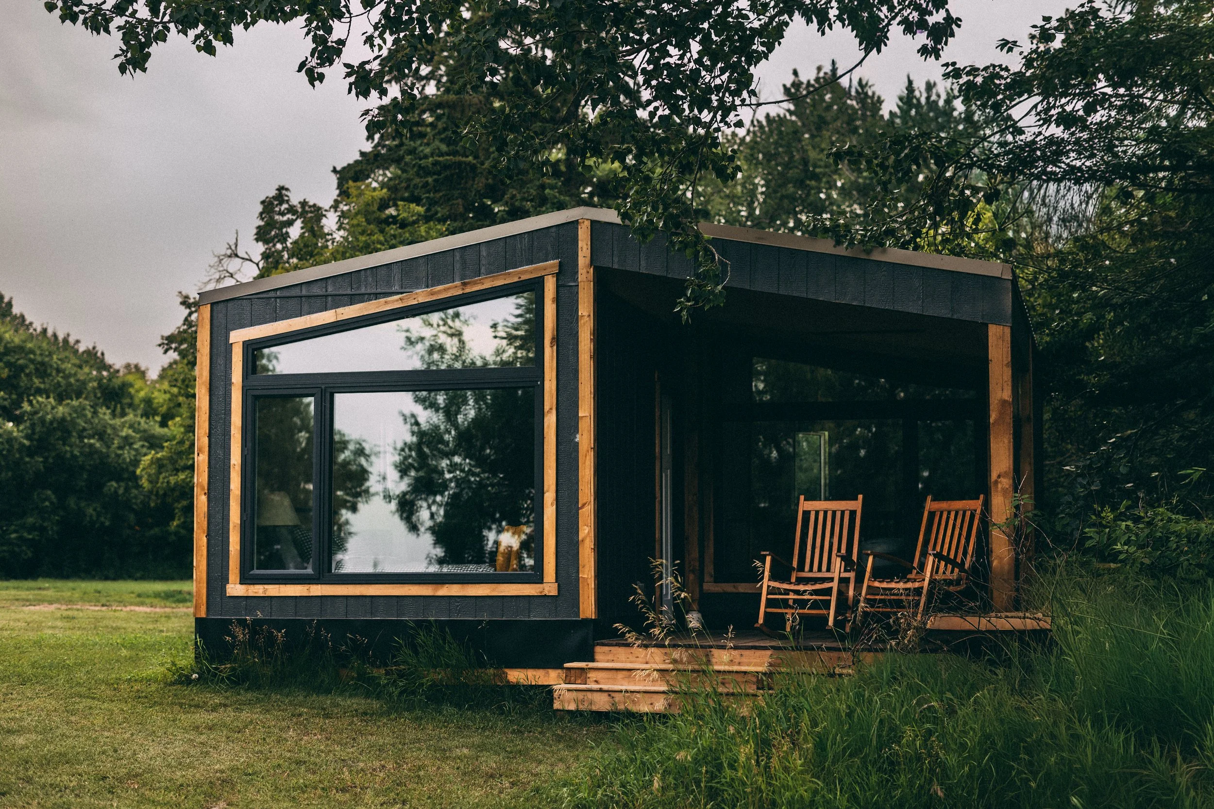 A modern tiny house with black exterior walls and large windows, surrounded by green trees and grass, with an outdoor wooden deck and chairs.