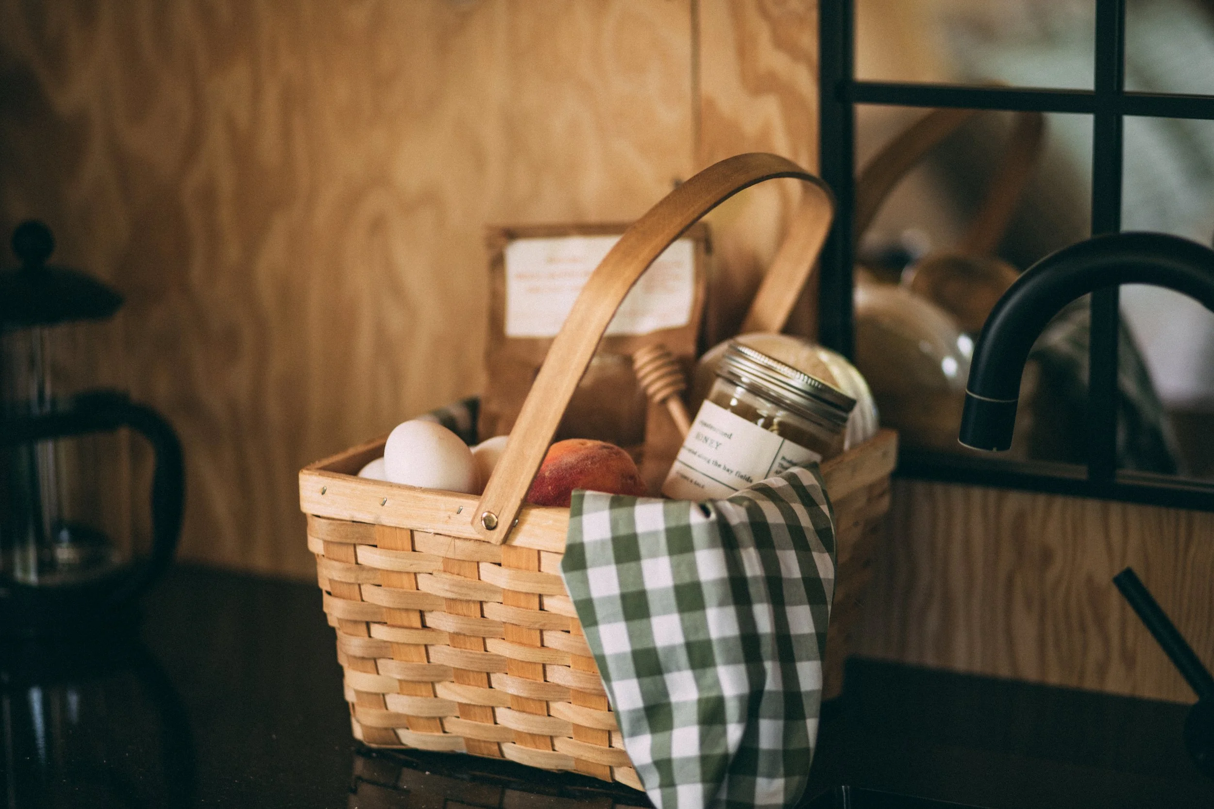 A wooden basket containing eggs, an apple, a jar of honey, and a piece of green checkered cloth sits on a black kitchen countertop.