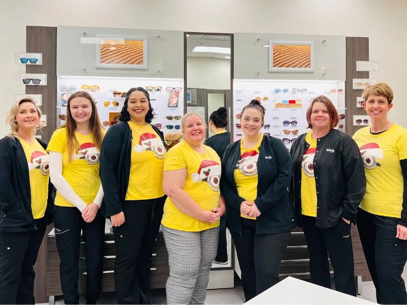 Group of six women standing inside an eyewear store, all wearing yellow shirts with cartoon eye designs and black jackets or pants, with sunglasses displayed on the wall behind them.
