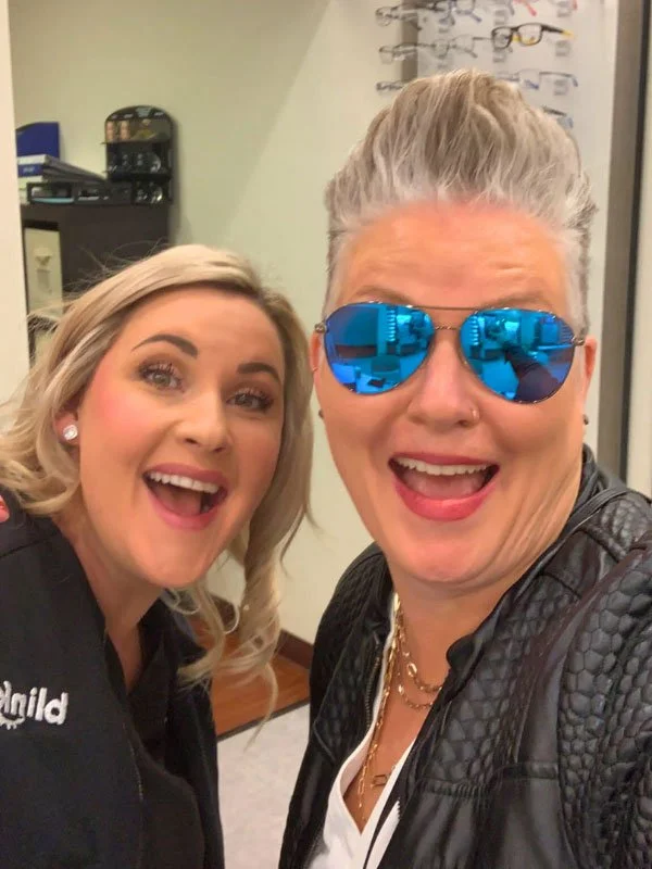 Two women smiling for a selfie indoors, one wearing reflective blue sunglasses and a black leather jacket, the other with blonde hair and earrings.