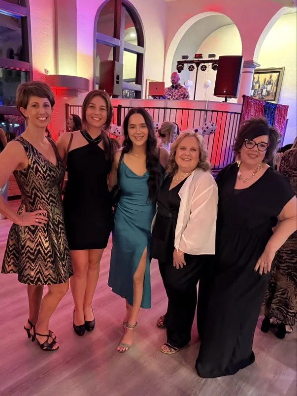 Five women in formal attire standing together at a celebration or party, with a man DJ in the background behind a railing.