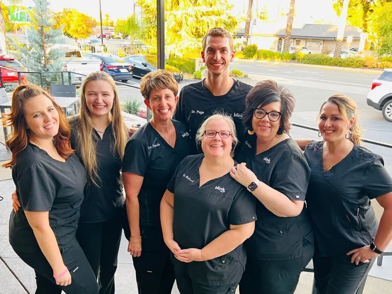 Six women in matching black medical scrubs posing together inside a building with large windows, overlooking a parking lot and trees outside.
