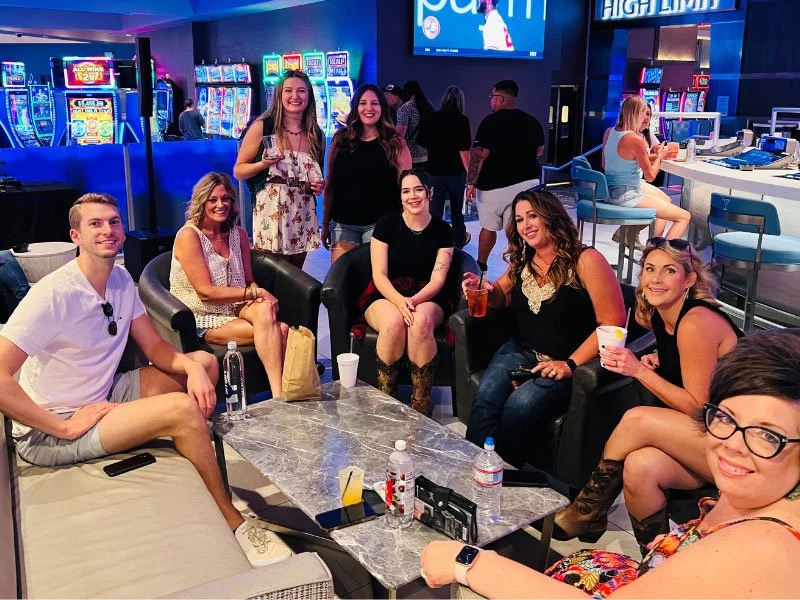 Group of seven friends sitting and standing in a casino lounge, with slot machines in the background, enjoying drinks and smiling at the camera.