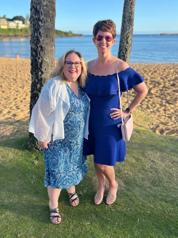 Two women standing on grass near the beach, smiling, with trees and water in the background. One woman wears a blue dress and sunglasses, the other a patterned dress with a white jacket and glasses.