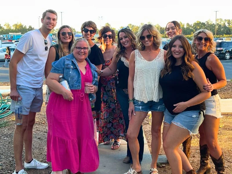 Ten young women and men posing outdoors in a parking lot, smiling and enjoying a sunny day.