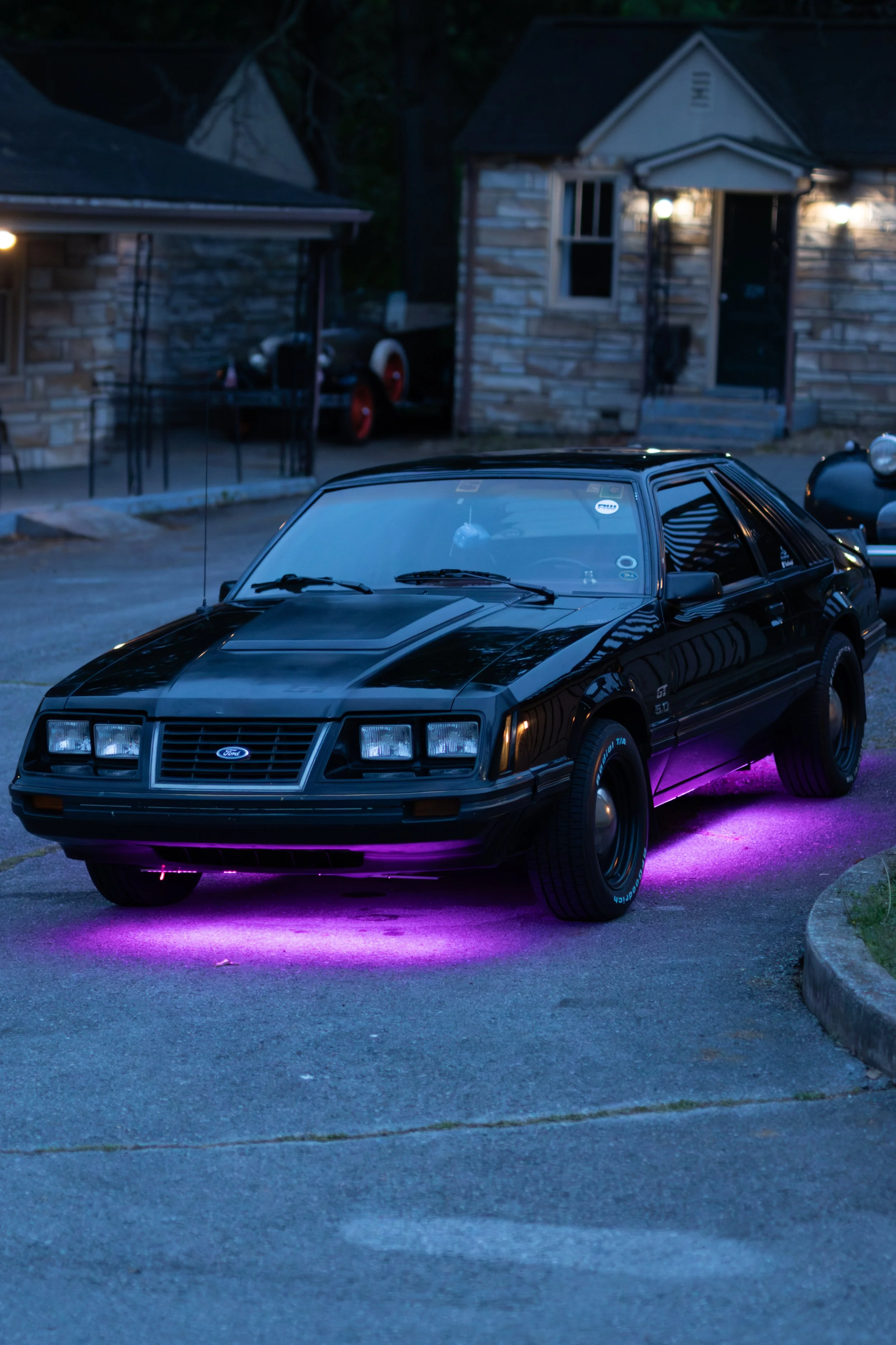 A black and gray vintage Ford Mustang GT 5.0 parked on grass at a car show during sunset, with other classic cars in the background.