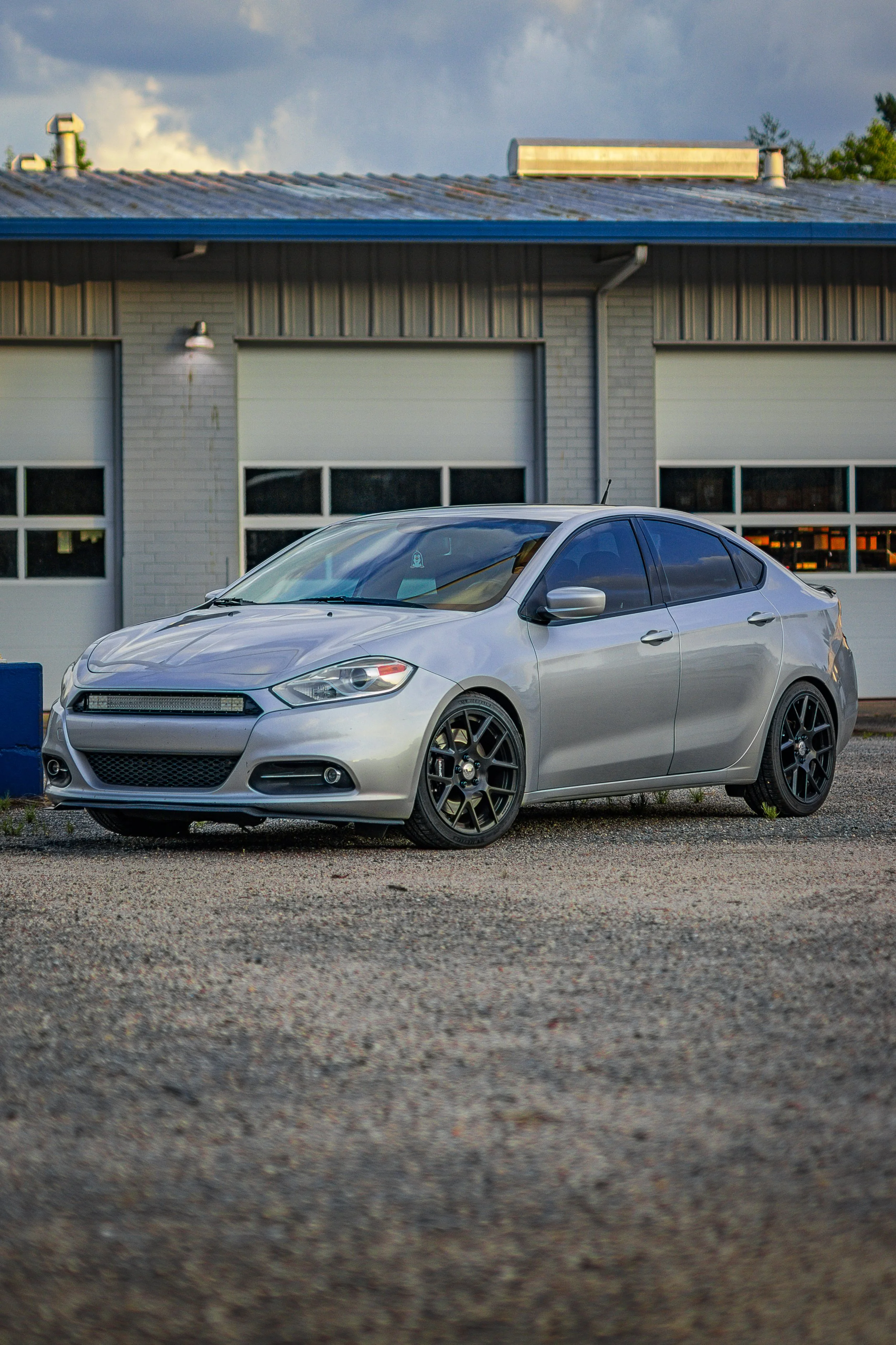 A silver car parked on a gravel area with trees and a body of water in the background during sunset.