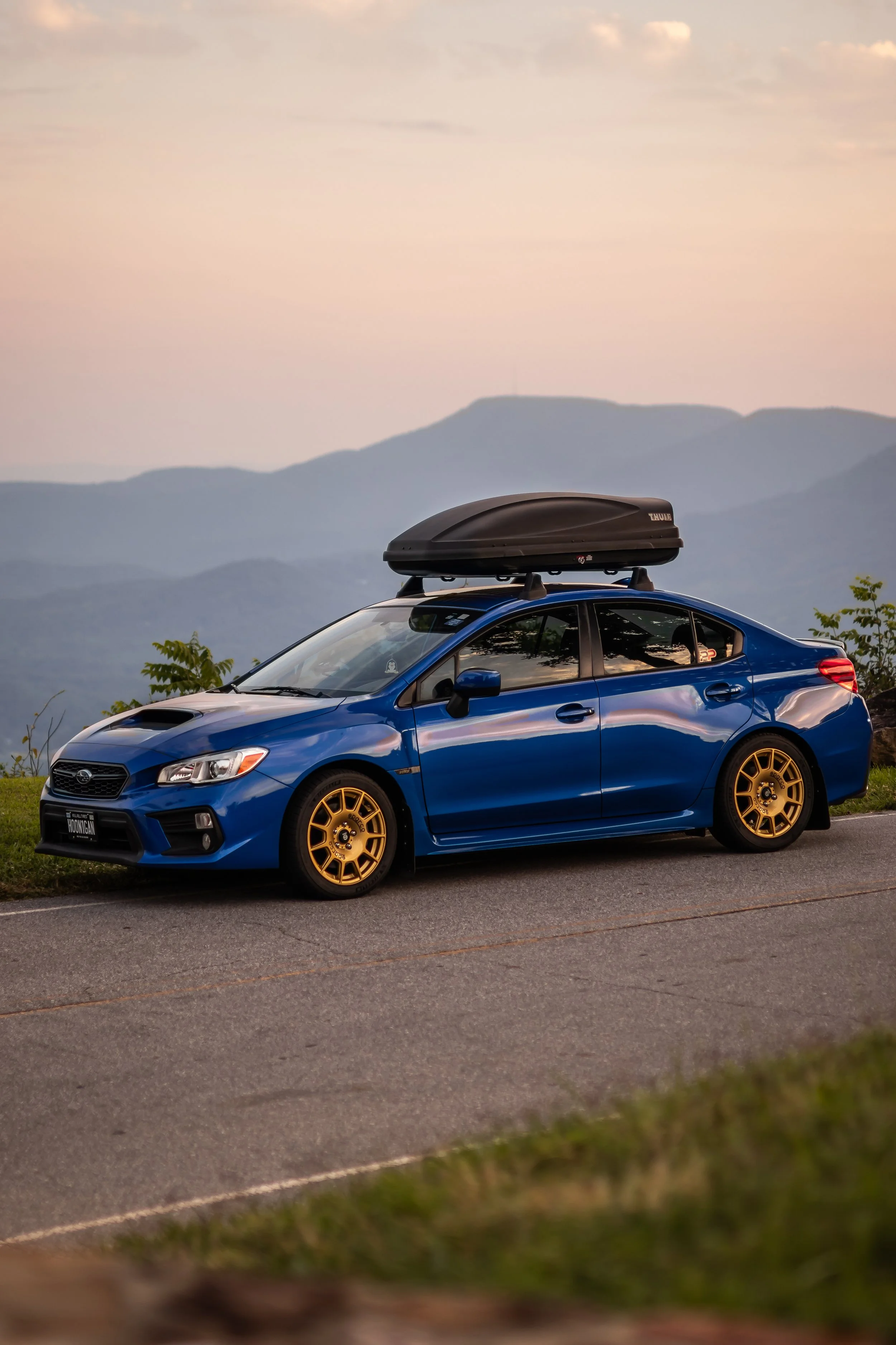 A blue Subaru WRX sedan parked in a parking lot at sunset with a black roof cargo box and gold wheels.