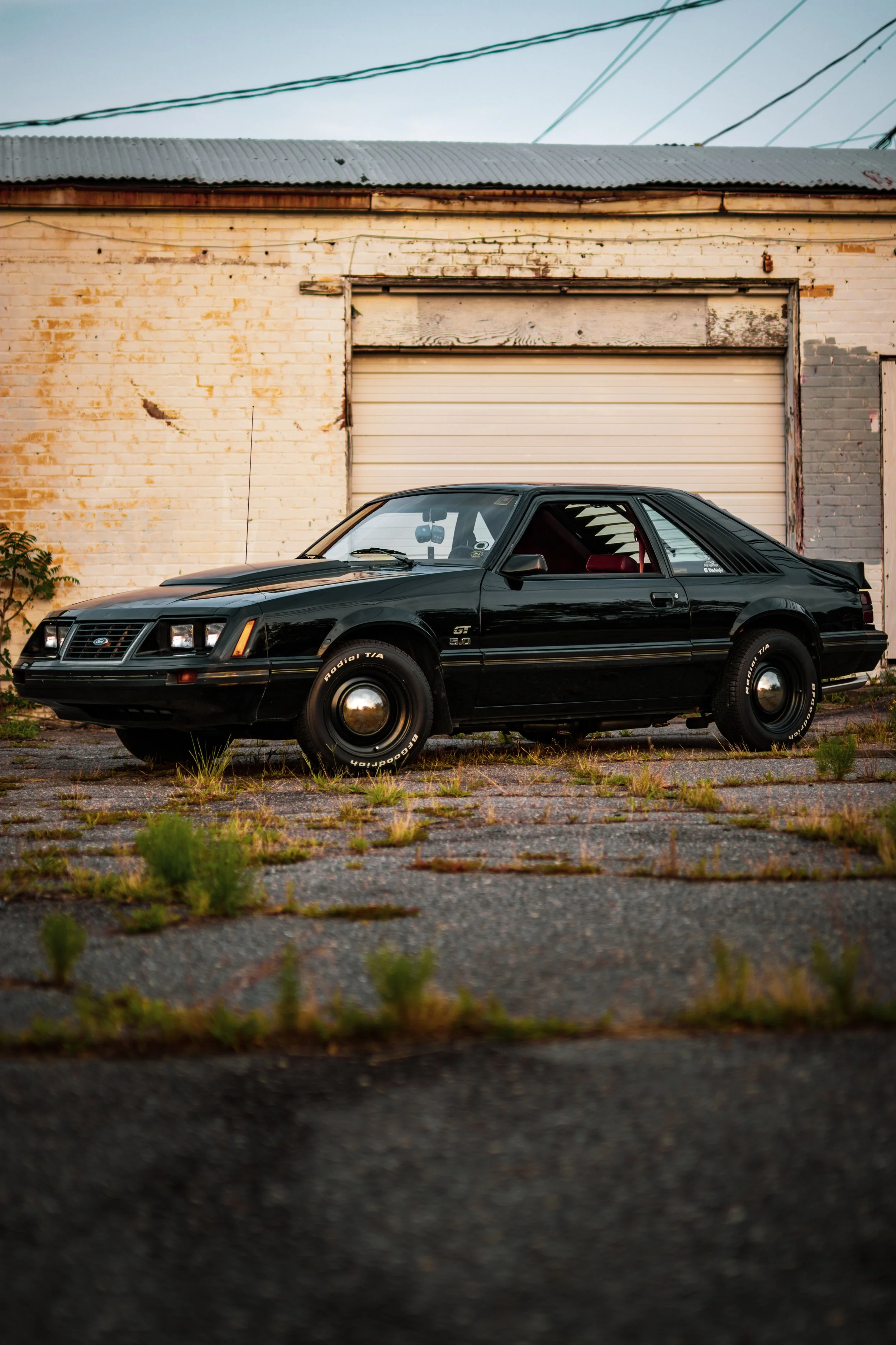 A black and gray vintage Ford Mustang GT 5.0 parked on grass at a car show during sunset, with other classic cars in the background.
