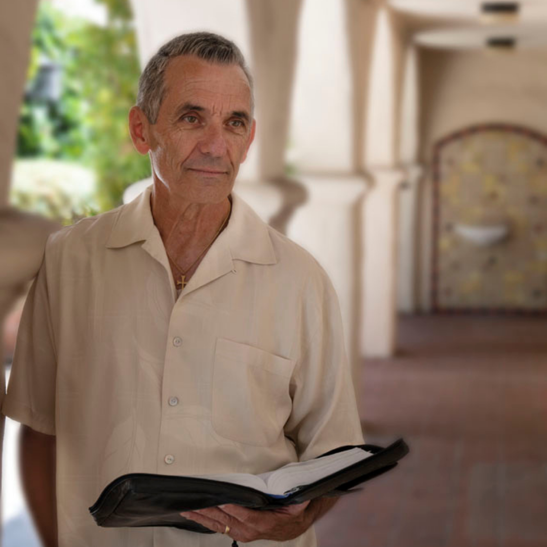 An older man with gray hair wearing a light beige short-sleeve button-up shirt holding an open black binder or book.