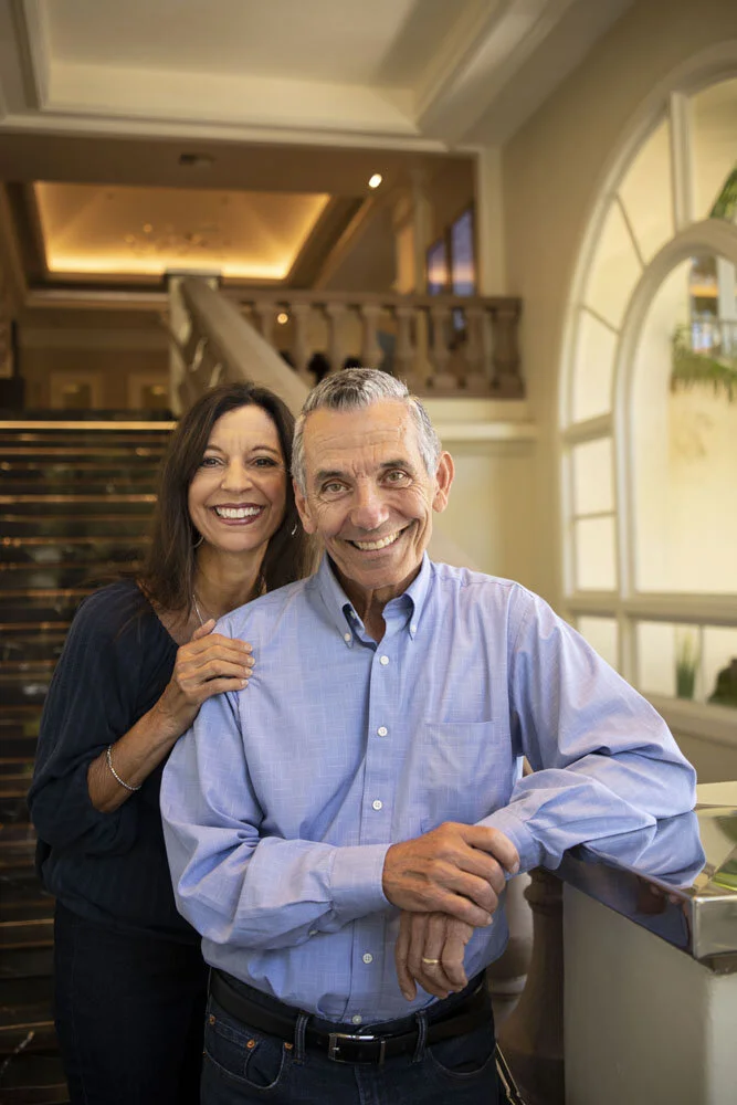 A smiling man and woman posing together indoors, with the woman standing behind the man and placing her hand on his shoulder. The setting appears to be a luxurious home with a staircase and large windows in the background.
