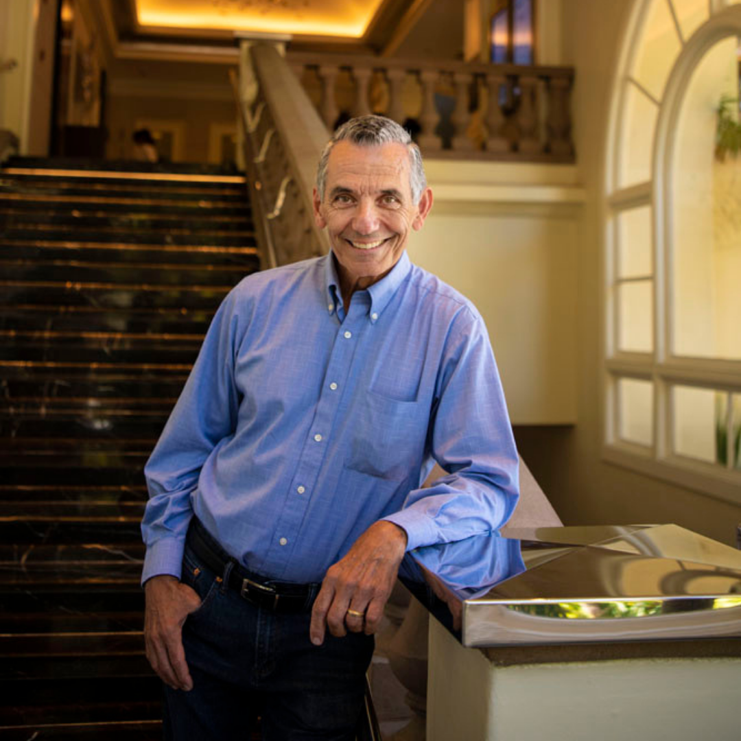 Smiling middle-aged man in a blue button-up shirt standing next to a counter in a well-lit, elegant interior with staircase and large windows.