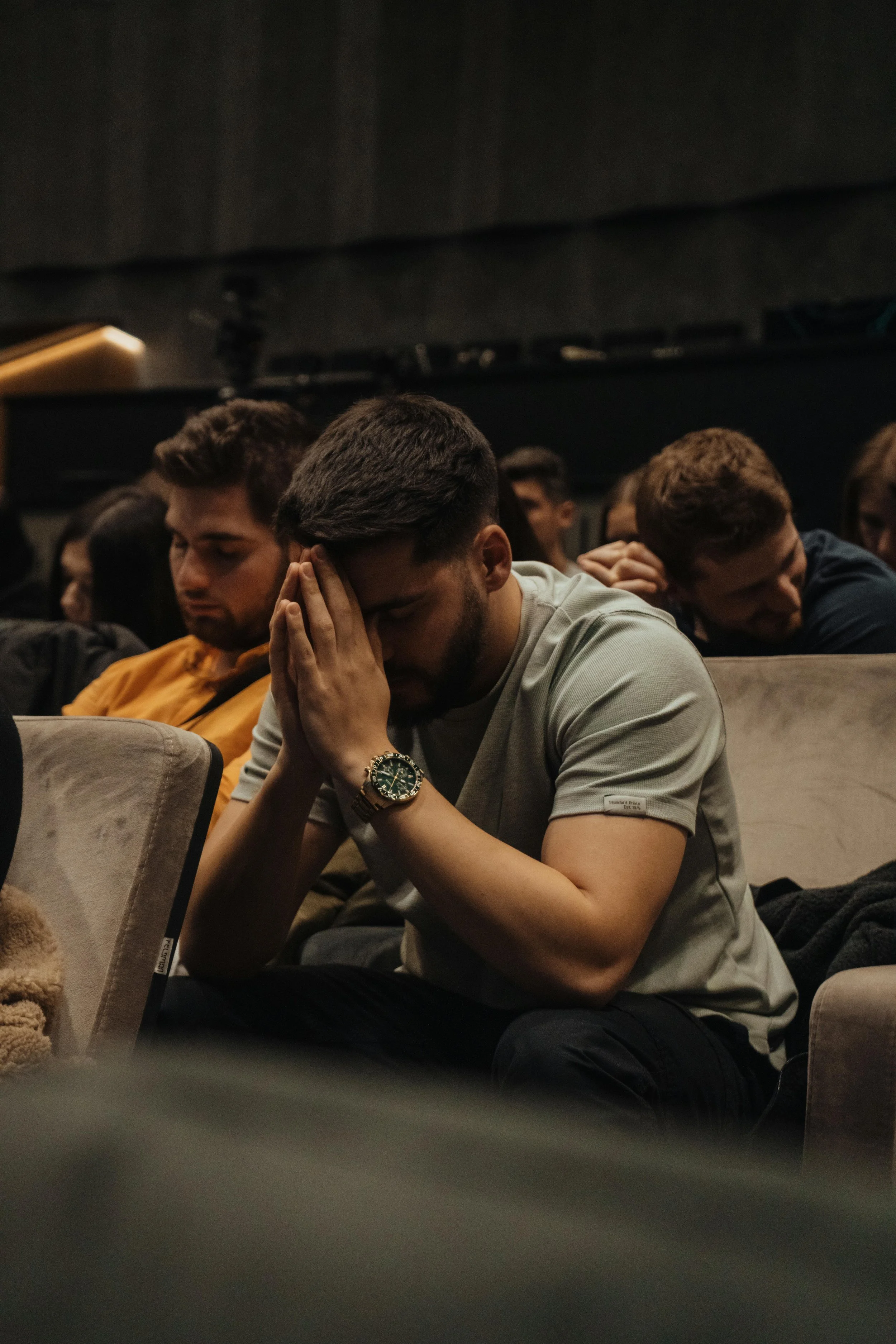 A group of young people sitting in an auditorium, some with their eyes closed and hands clasped, appearing to be in prayer or reflection.