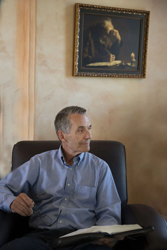 An older man seated in a black leather chair, holding glasses and an open book, with a painting hanging on the wall behind him.