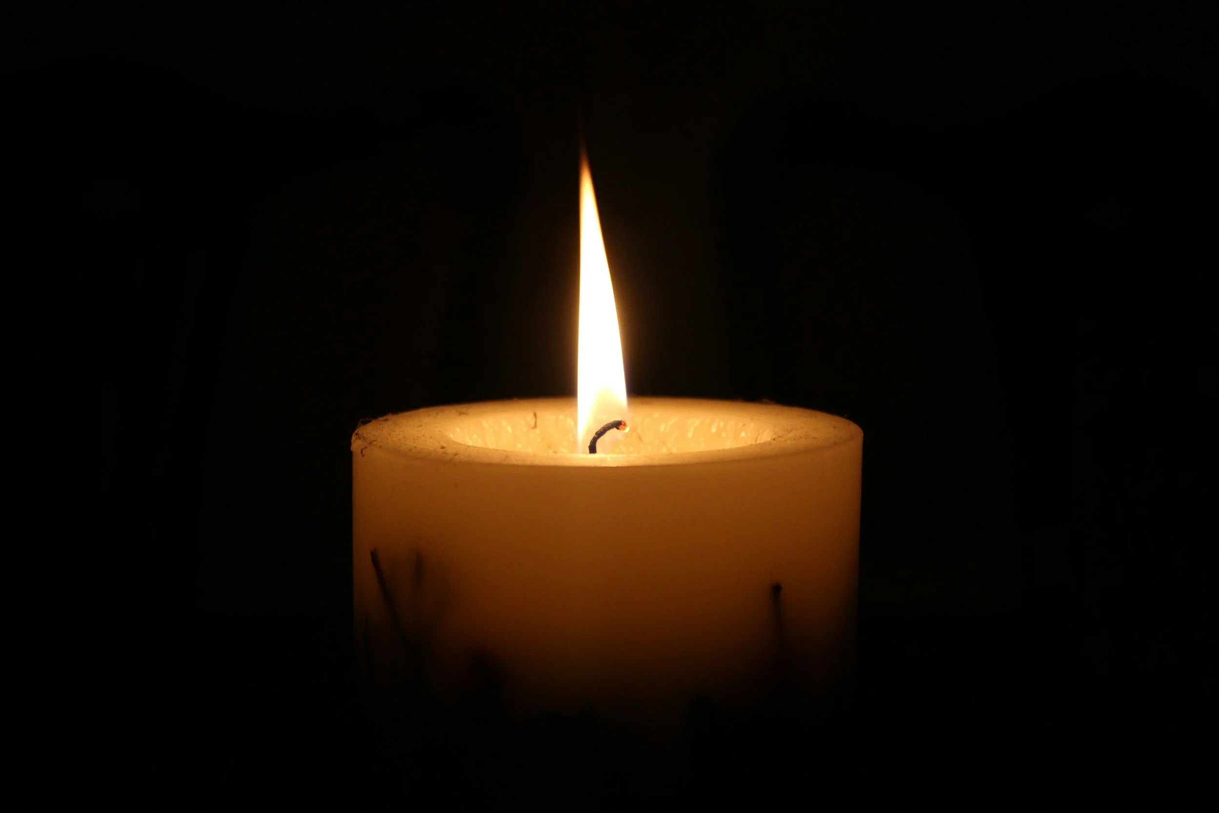 Close-up of a burning candle with a tall, steady flame, set against a dark background.