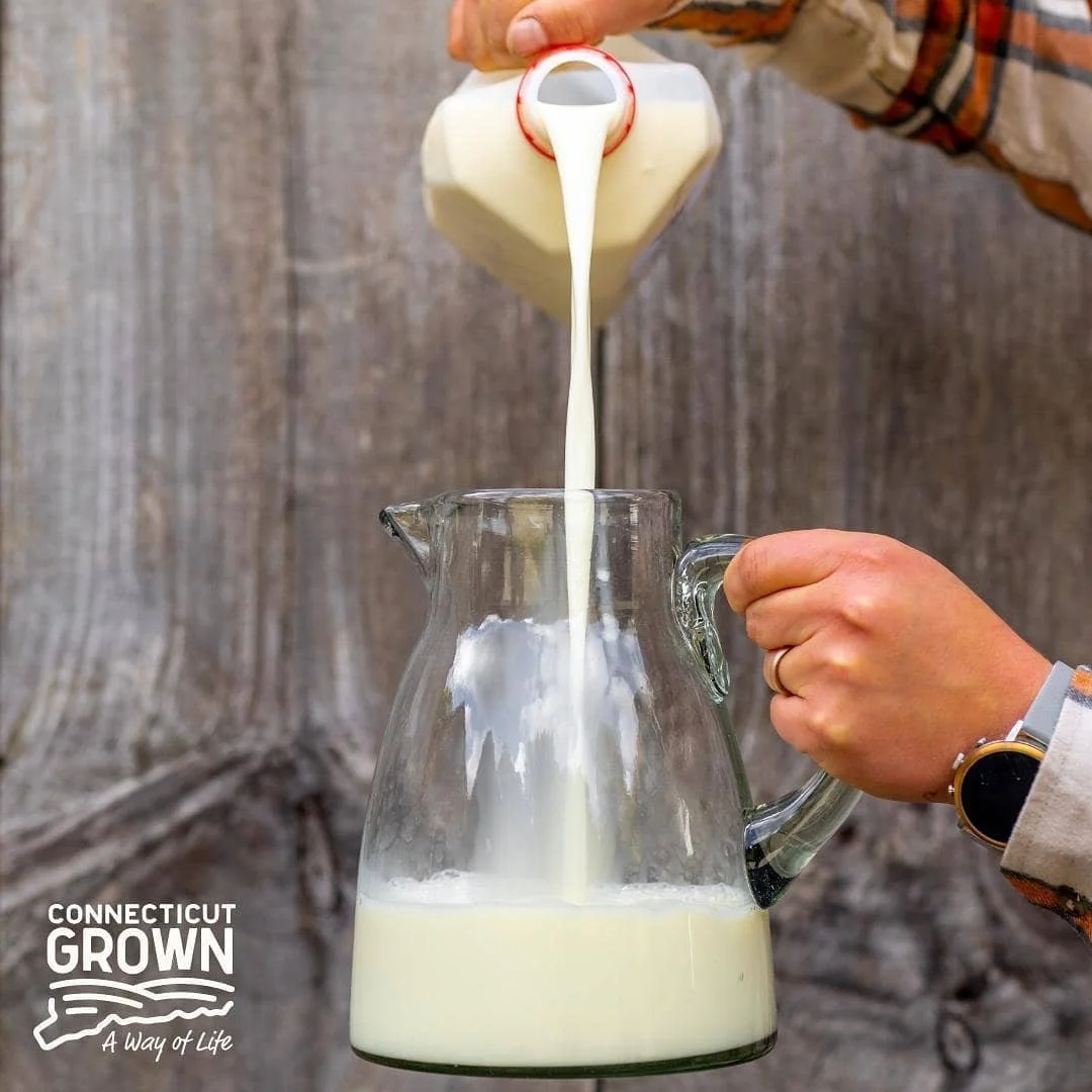Person pouring milk into a glass pitcher, with a wooden background and a Connecticut Grown logo in the corner.
