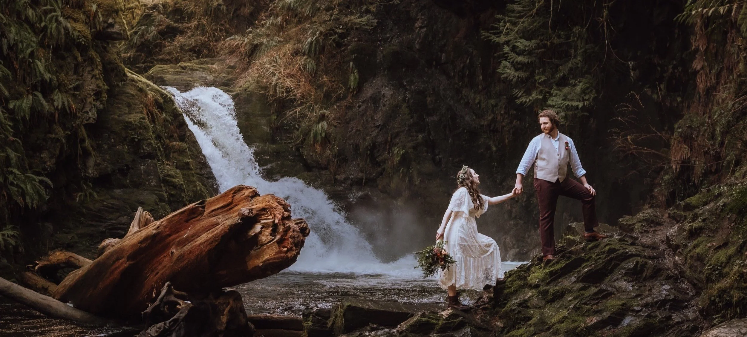 A couple getting married outdoors in a forest by a waterfall, with the woman in a white dress holding a bouquet and the man in dress pants and a vest, holding her hand.