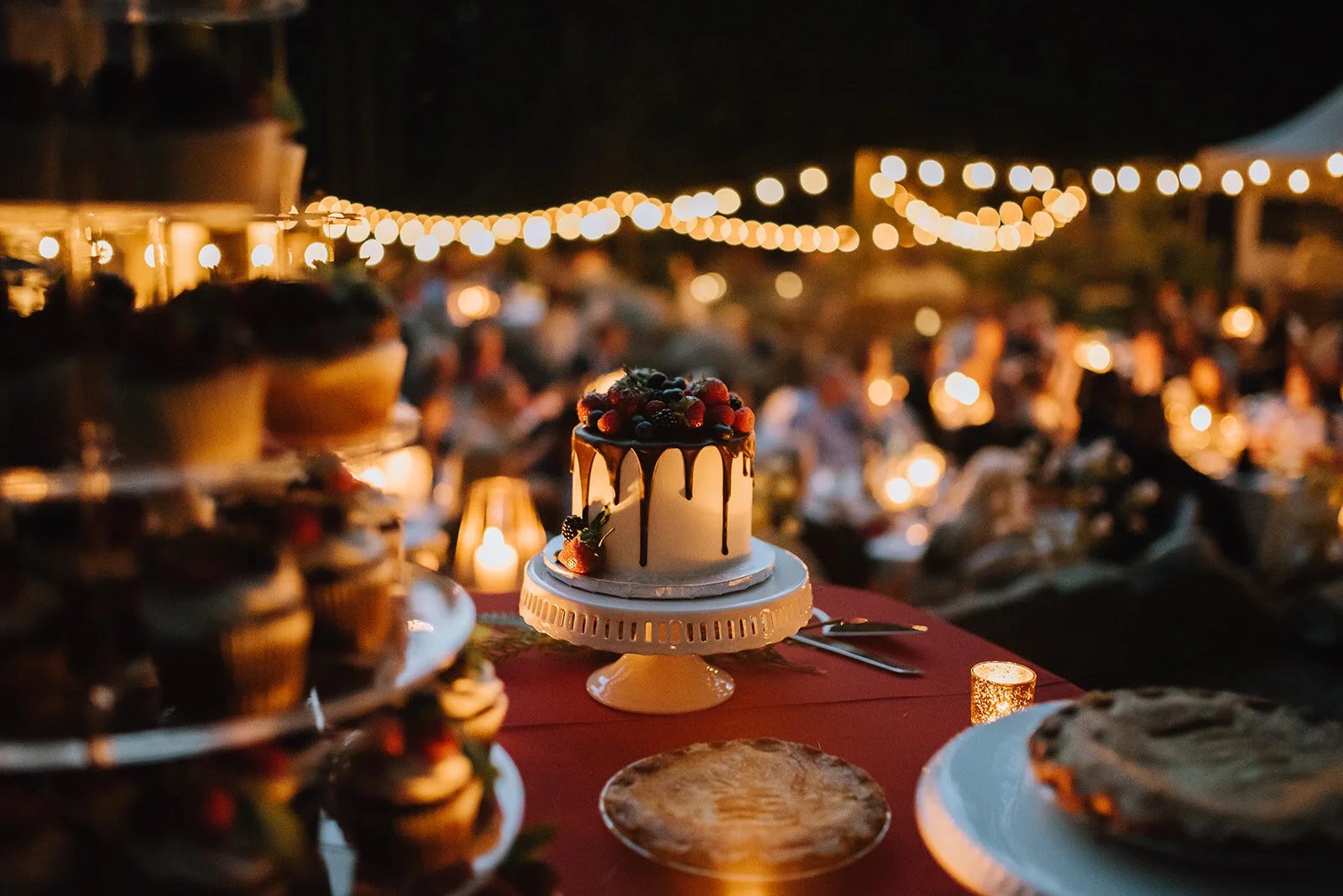 A birthday cake with fruit on top is on a table at an outdoor celebration, with string lights and a blurred crowd in the background.