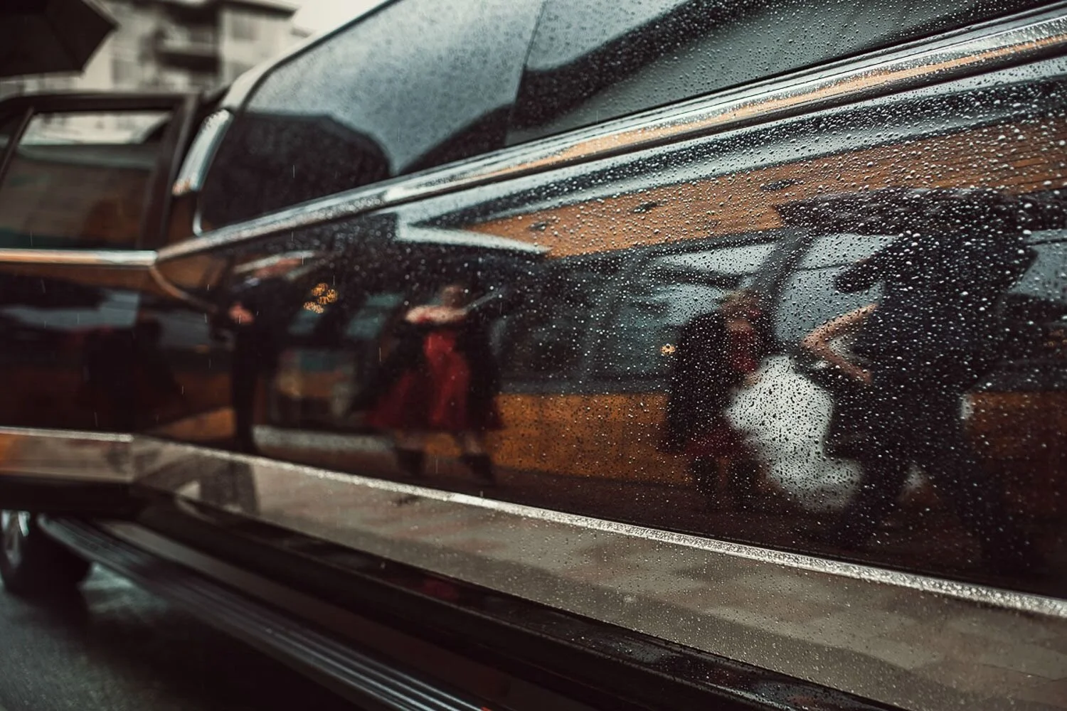Close-up of a black car with rain droplets on its surface, reflecting nearby buildings and people.