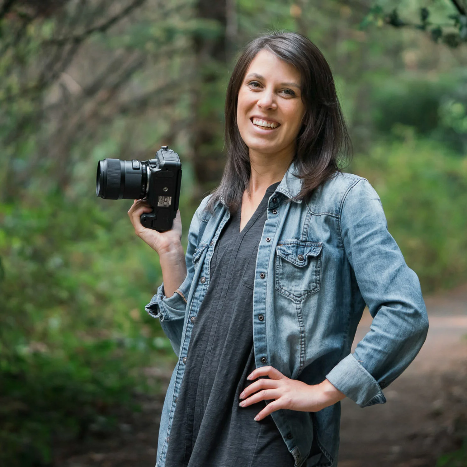 A woman outdoors on a trail holding a camera with a forested background, smiling at the camera.