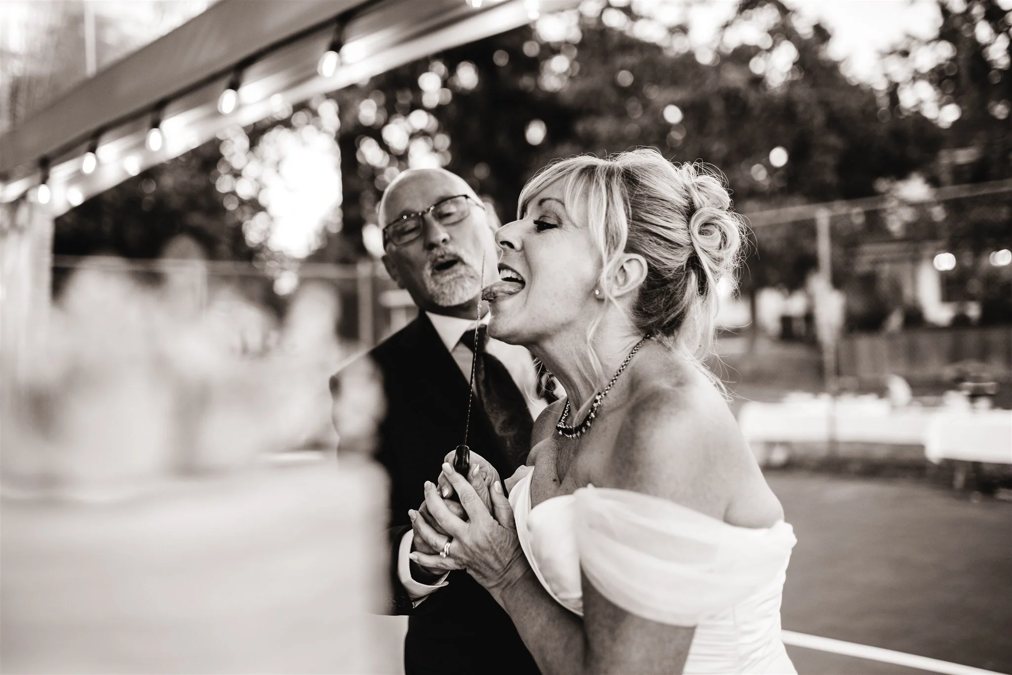 A black-and-white photo of a woman in a wedding dress and a man in formal attire playing with a lollipop while standing outdoors.