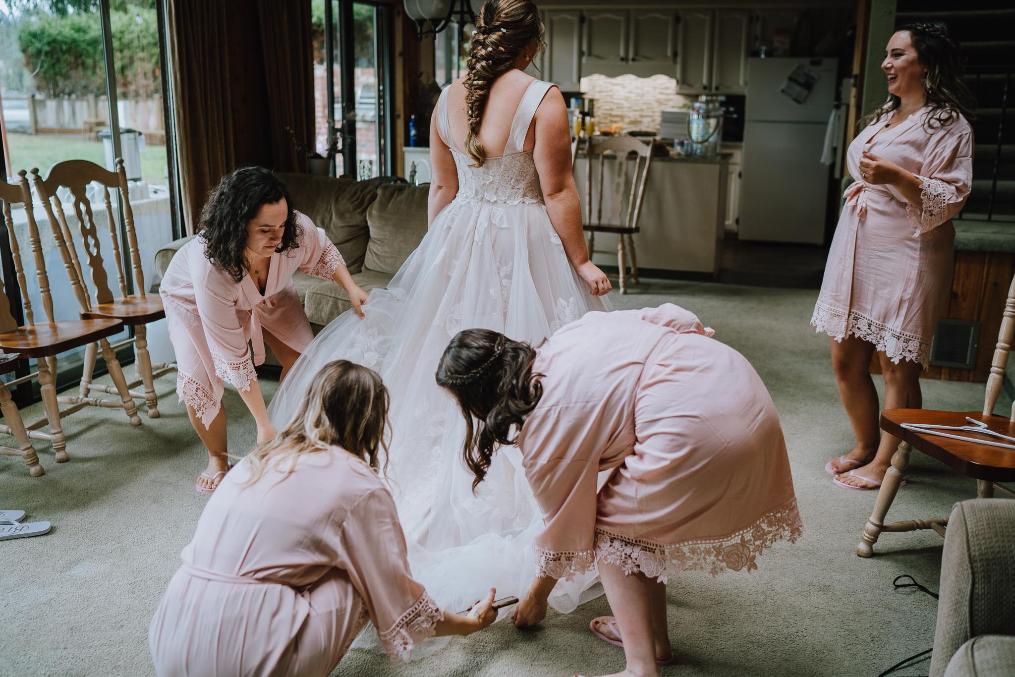 Women preparing a bride in a white wedding gown inside a cozy living room.