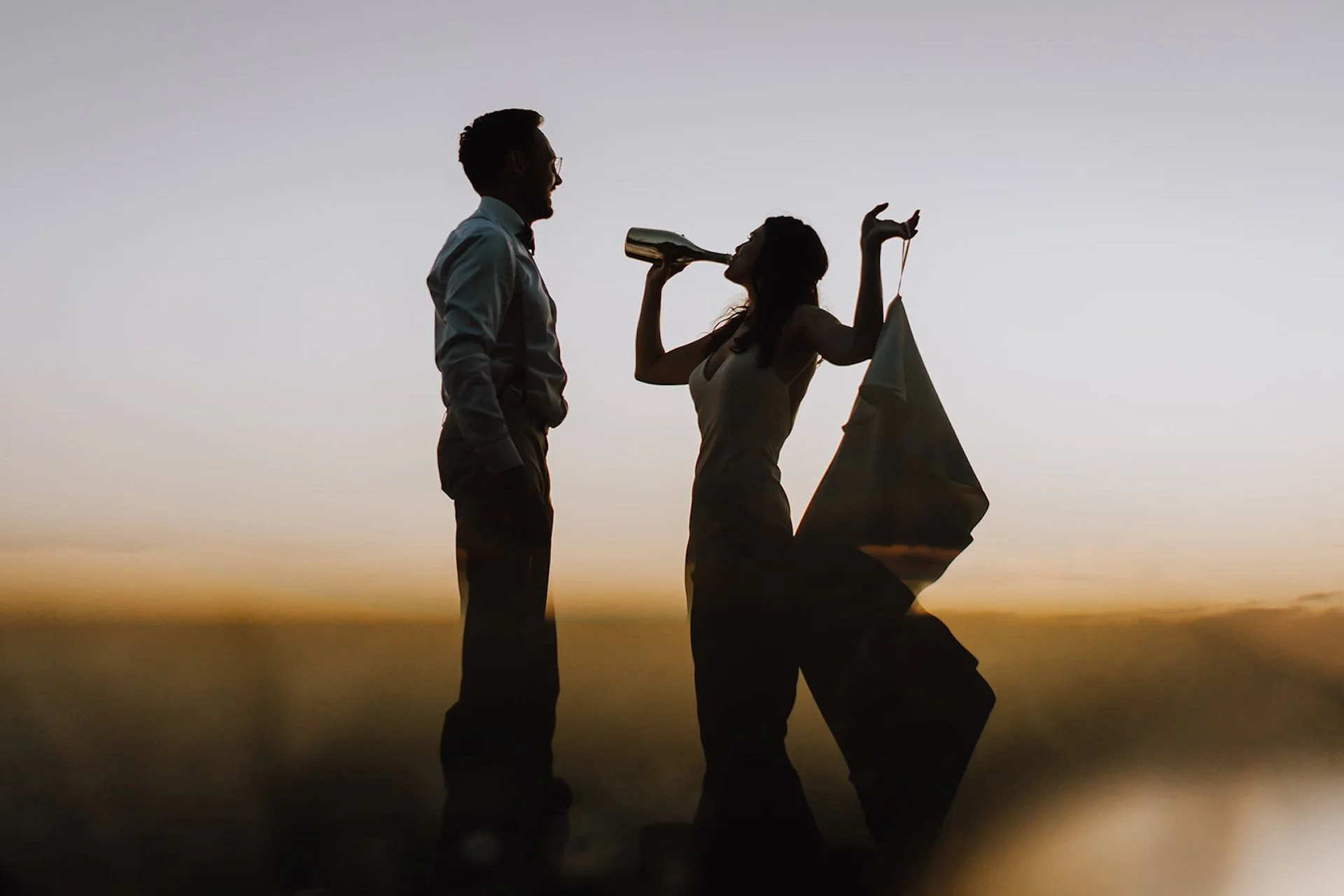 Silhouette of a couple at sunset, with the woman holding a flag and the man drinking from a champagne bottle.
