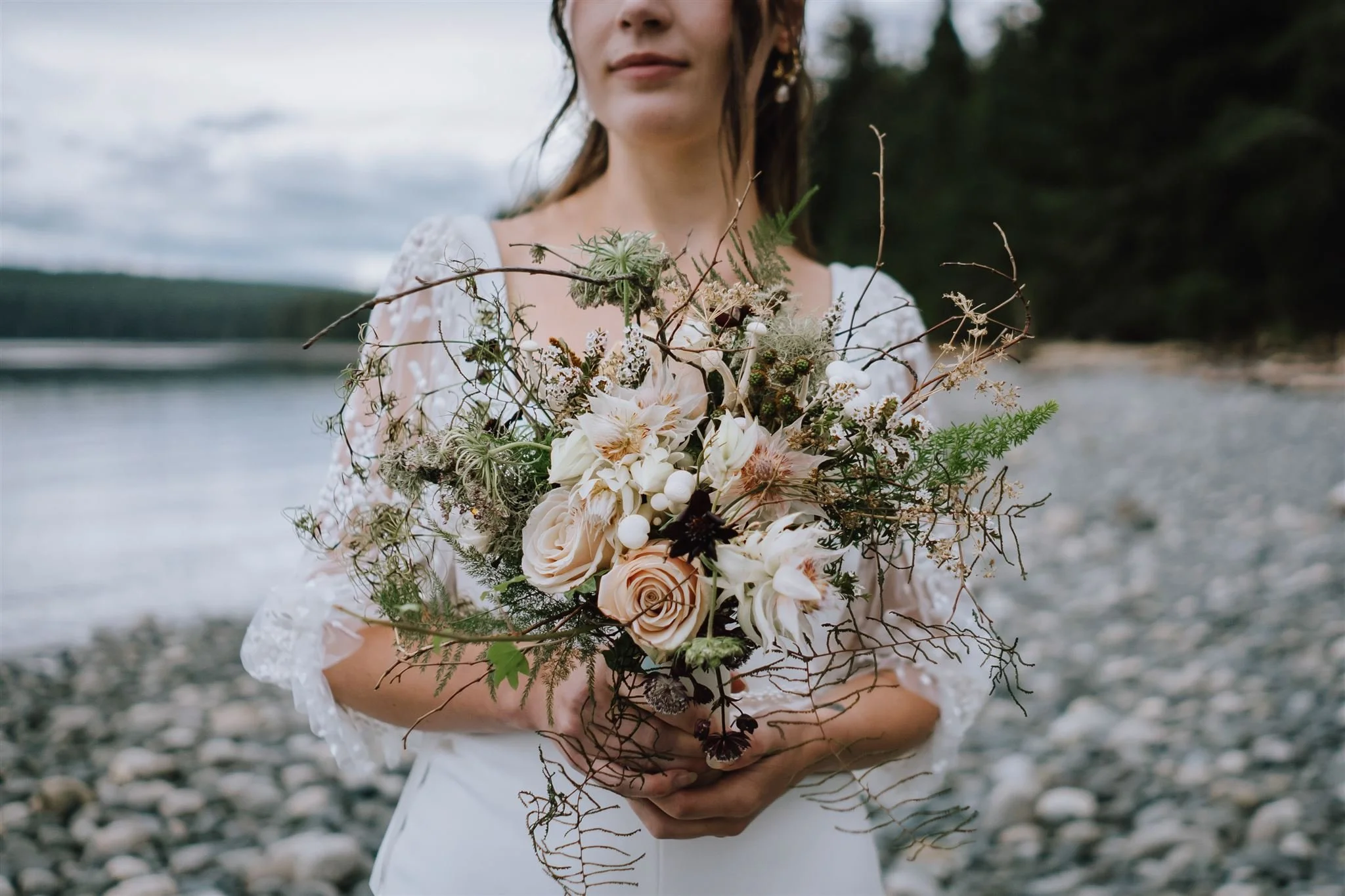 A woman in a white dress holding a bouquet of flowers near a river or lake with a rocky shoreline and trees in the background.