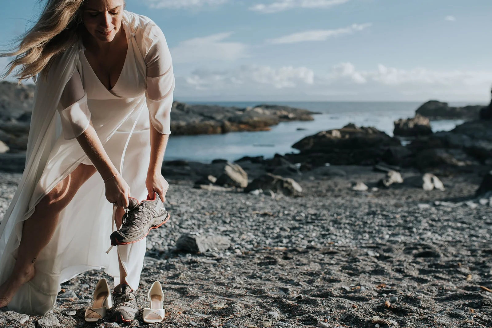 A woman in a white dress is bent over on a rocky beach, putting on a sneaker. A pair of high-heeled shoes are on the ground beside her. The ocean and rocks are visible in the background with a partly cloudy sky.