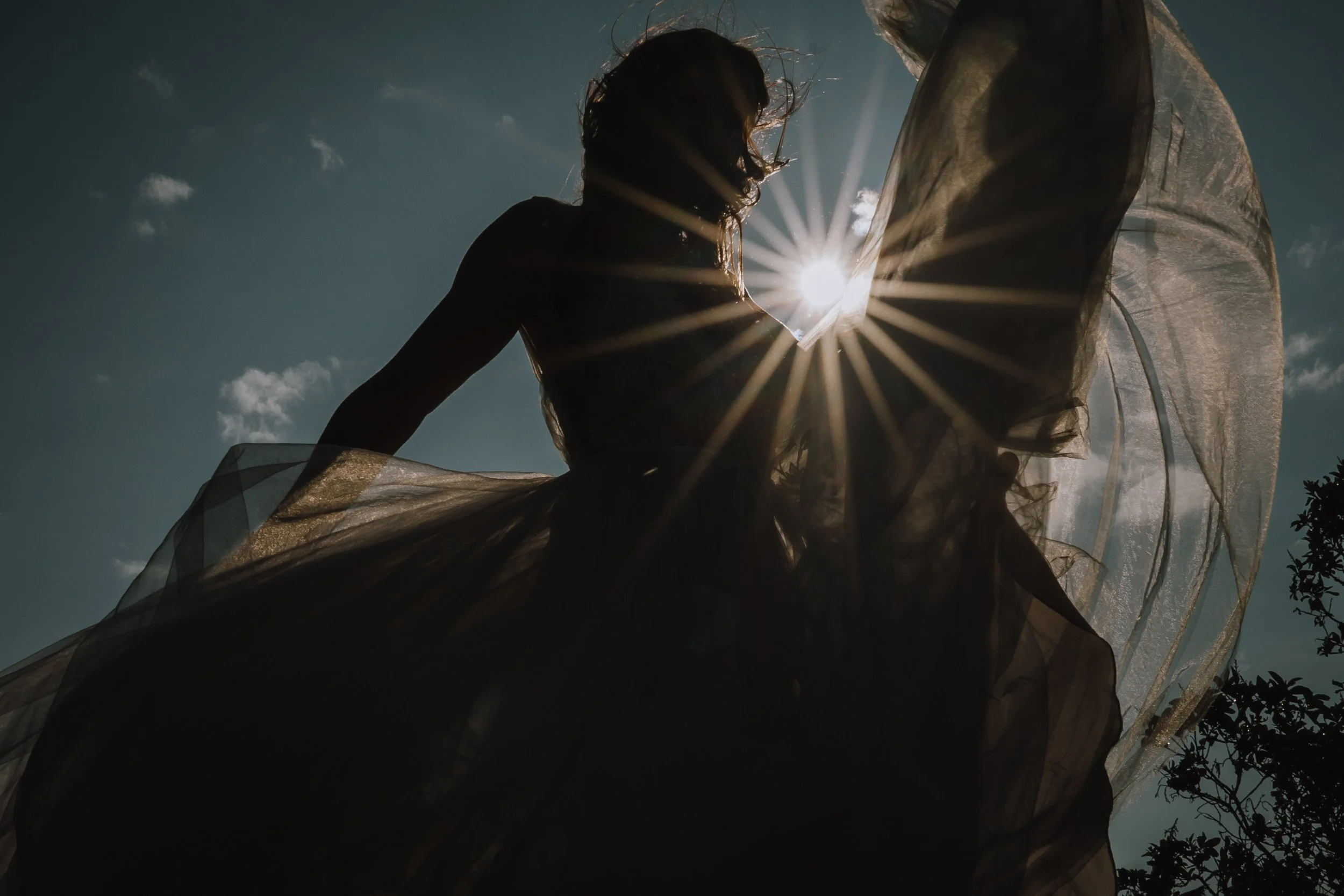 Silhouette of a woman with flowing fabric, backlit by the sun with rays radiating outward, against a blue sky with a few clouds.