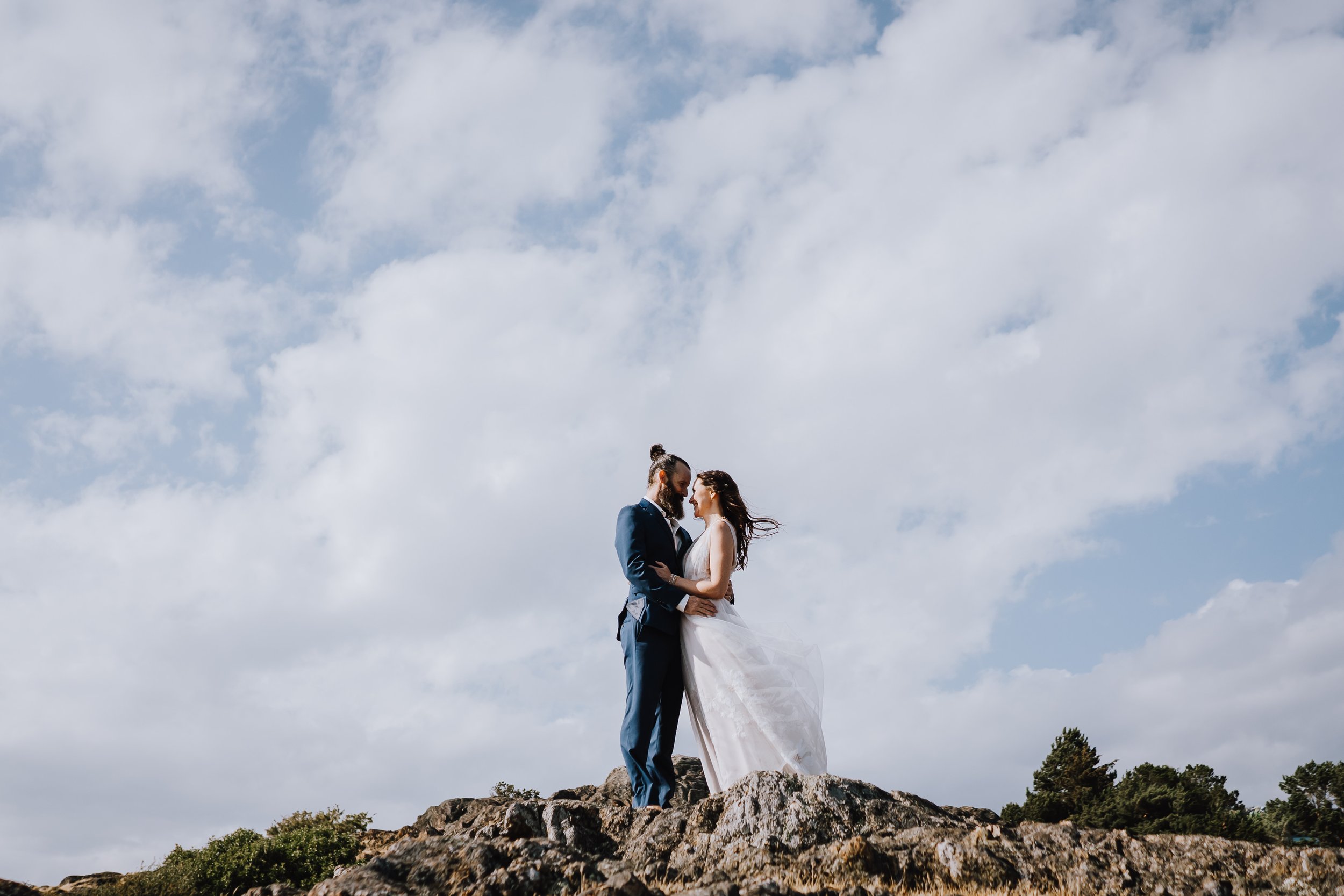 A bride and groom standing on rocks outdoors, embracing, with cloudy sky and trees in the background.