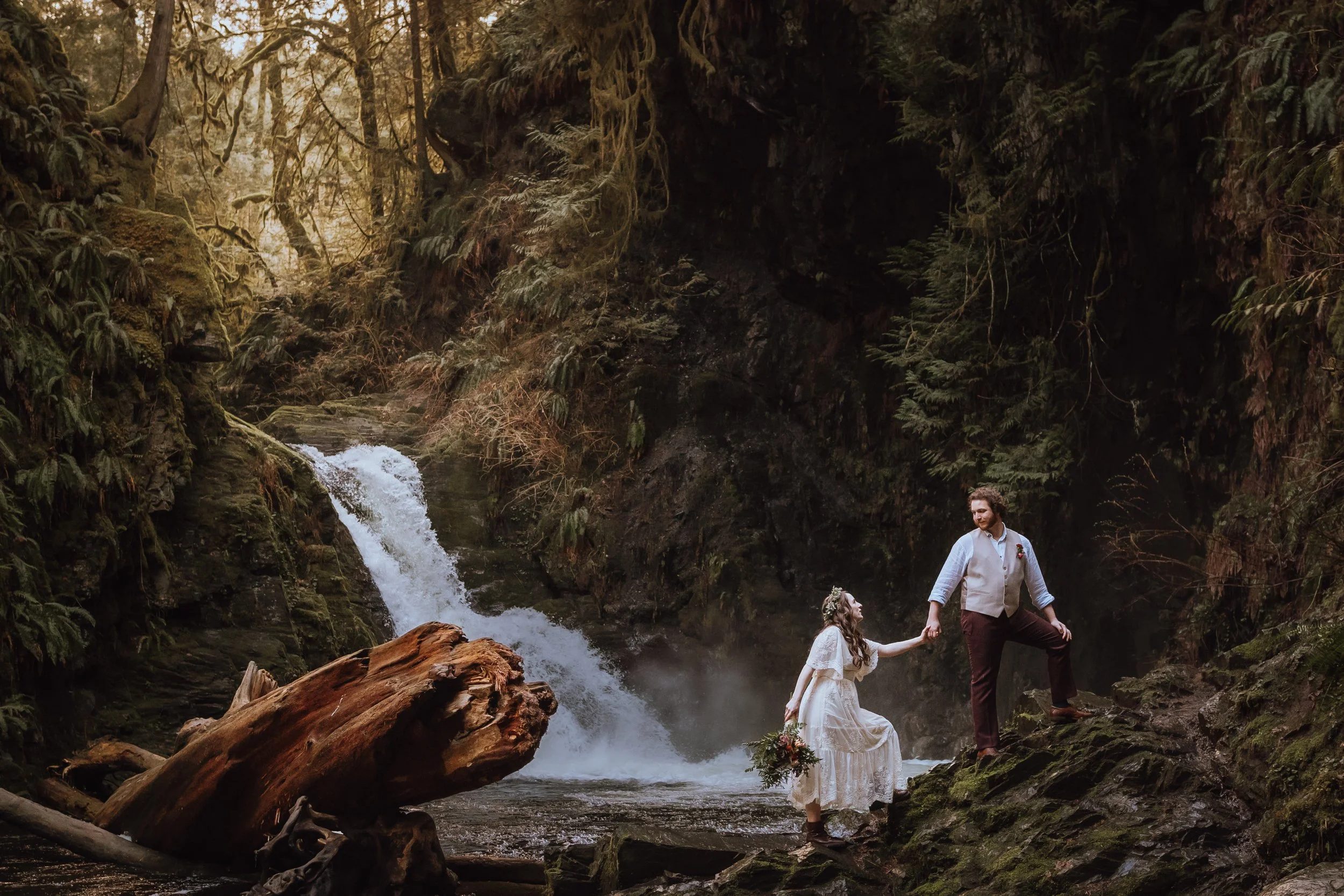 A couple in wedding attire standing near a waterfall in a forest, holding hands, with greenery and moss-covered rocks surrounding them.