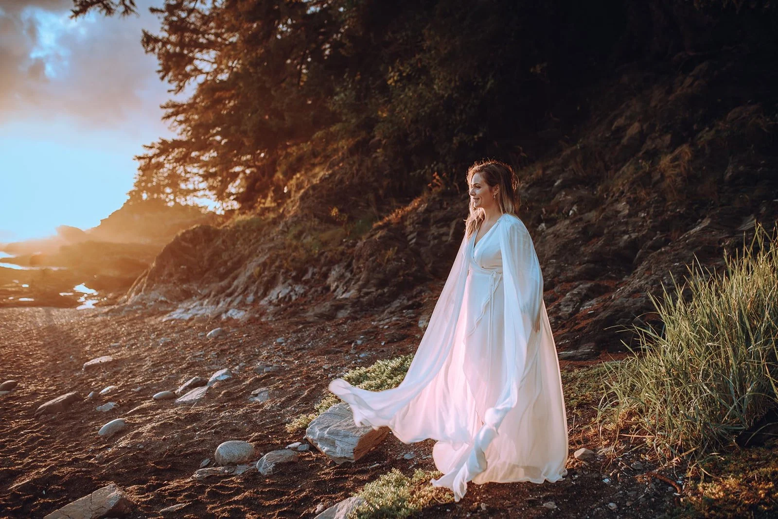 A woman in a white flowing dress stands on a rocky beach with grass, during sunset, with trees and cliffs in the background.