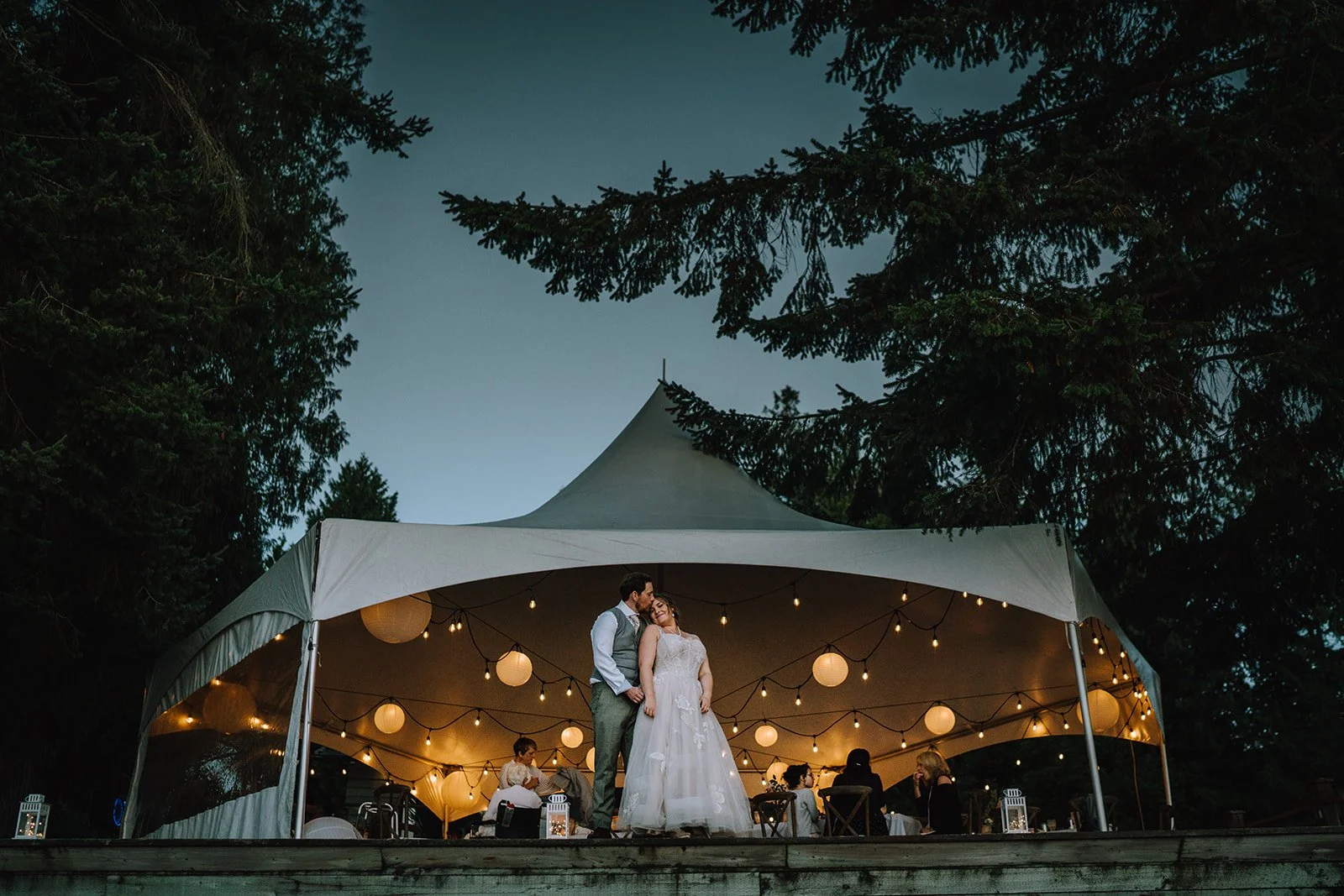 A couple dances under a large white tent decorated with string lights and paper lanterns at dusk or evening, with trees in the background.