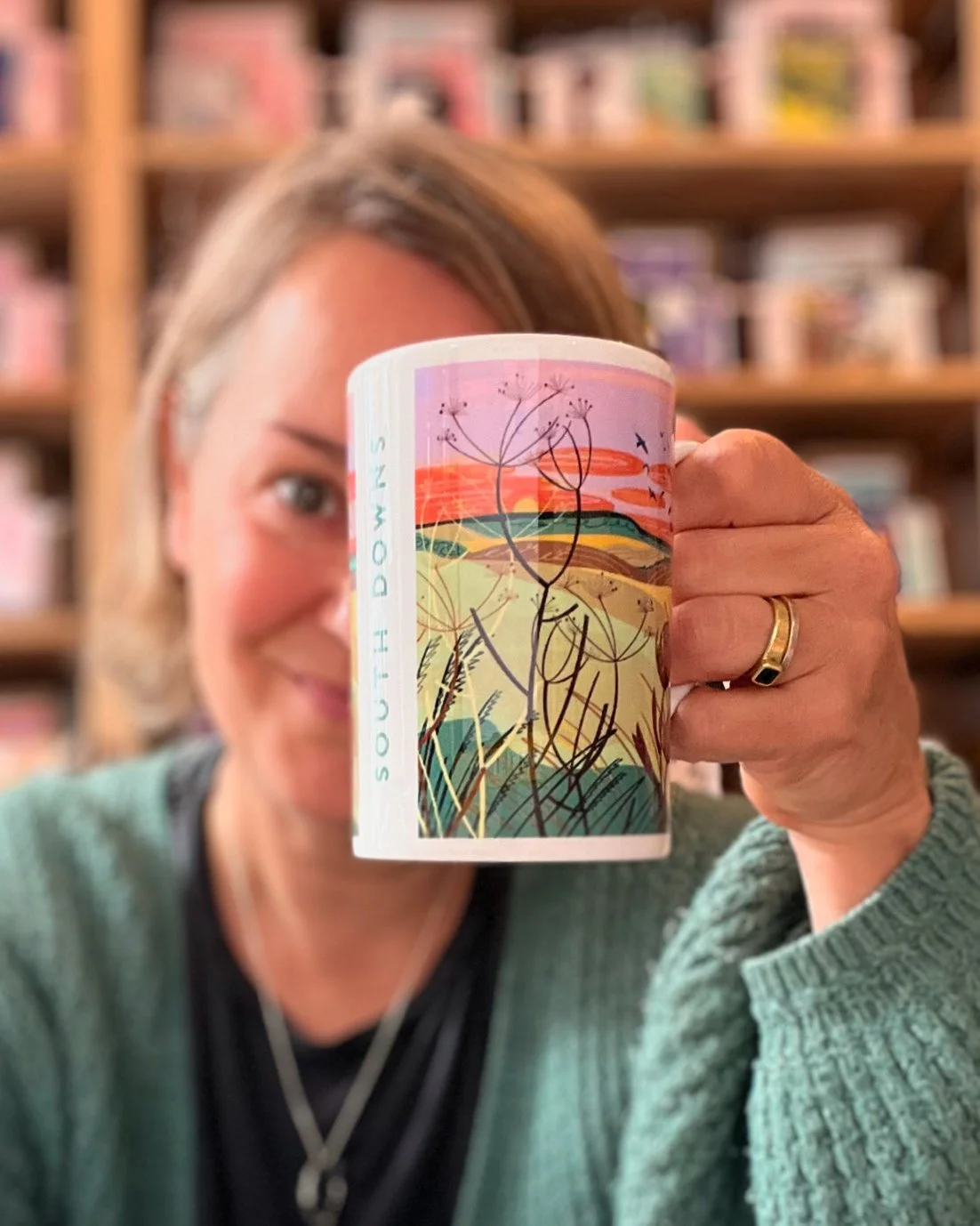 A woman holding a colorful coffee mug with landscape and plants design in front of her face, blurred bookshelf background.