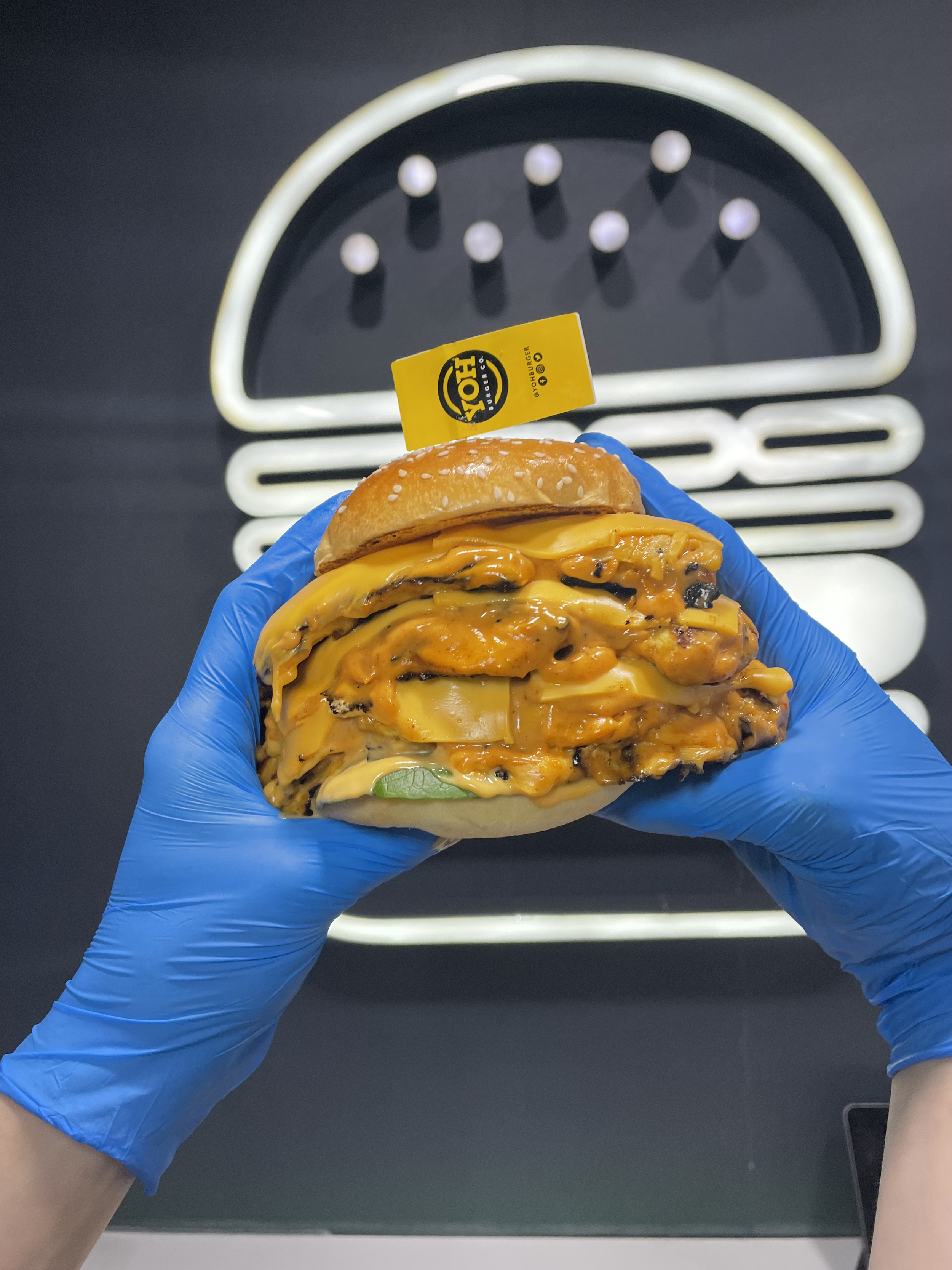 Person wearing blue gloves holding an oversized cheeseburger with melted cheese and toppings in front of a black and white decorative background.